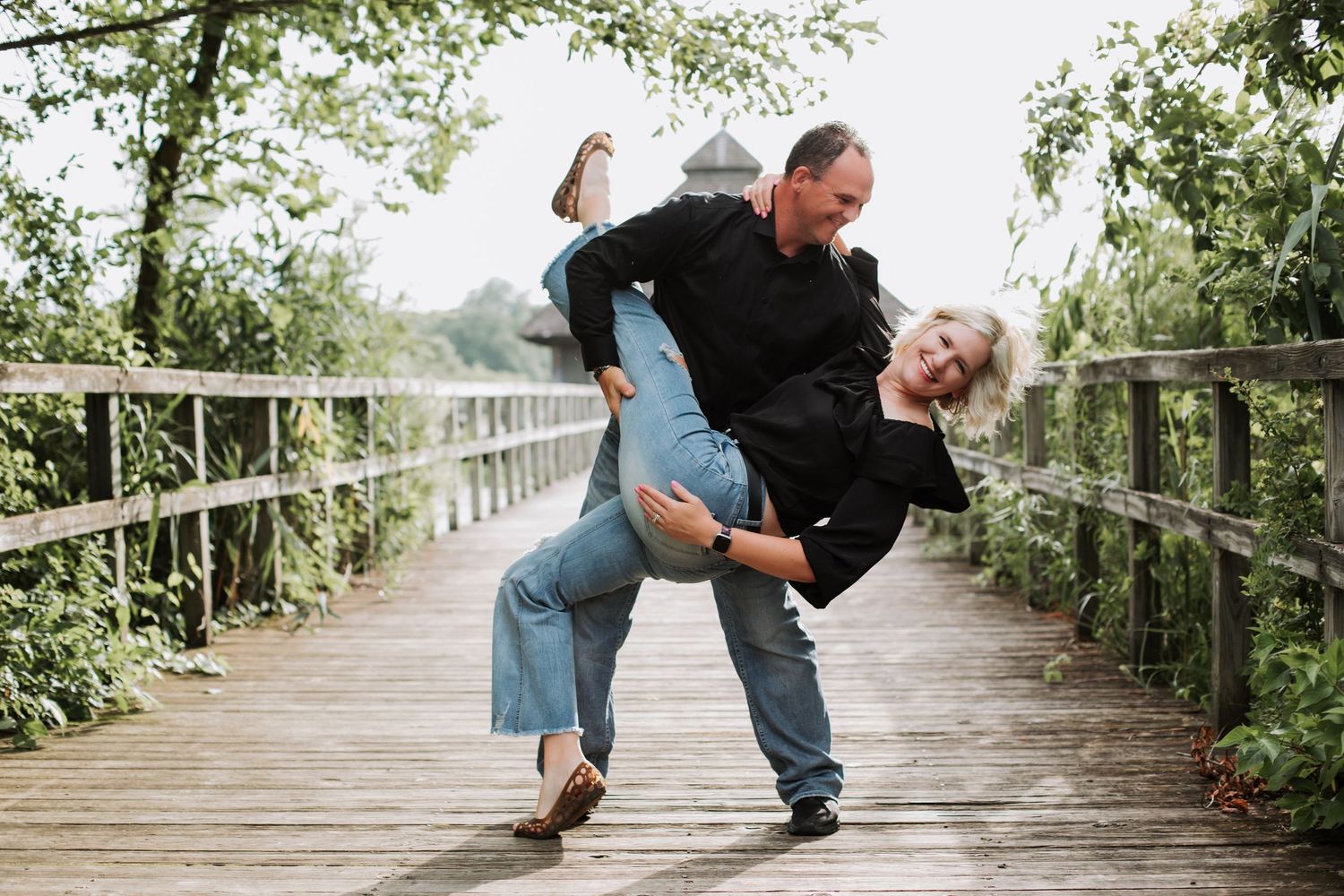 Man playfully dipping woman on a wooden bridge surrounded by green trees.