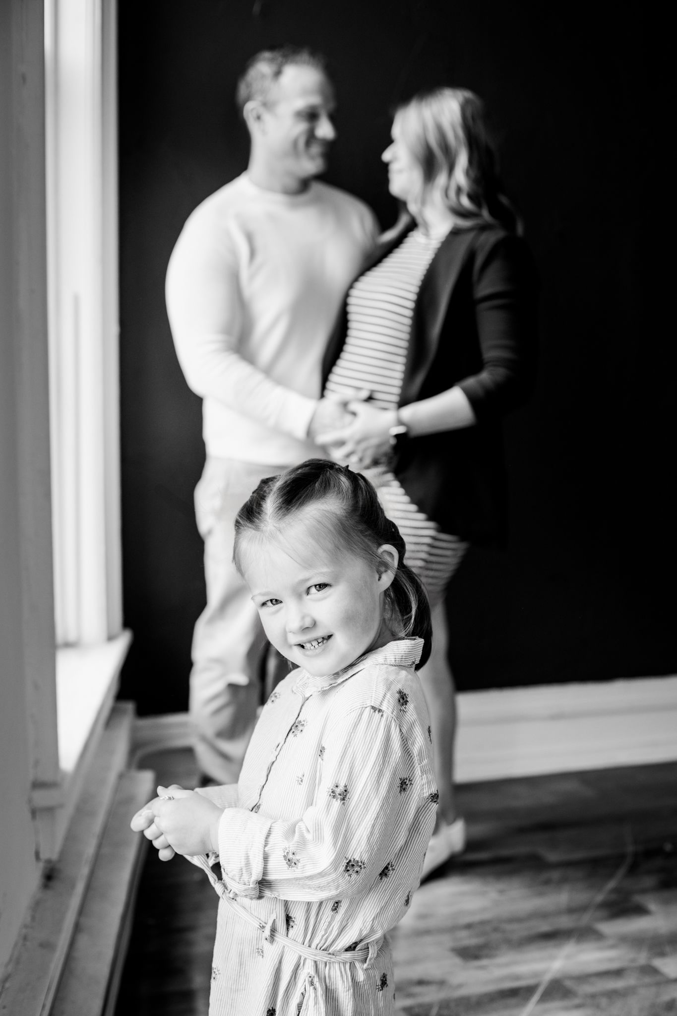 Young girl smiles at camera while parents hold hands and embrace in black and white family portrait.
