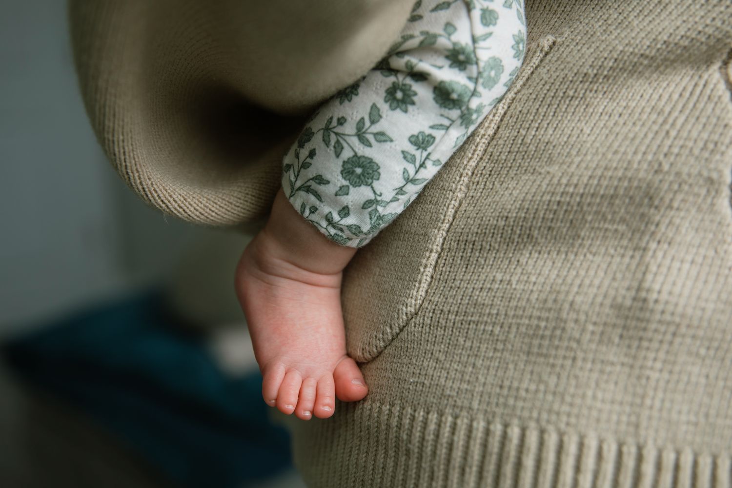 Close-up of newborn baby foot peeking from floral onesie beside beige knit sweater.