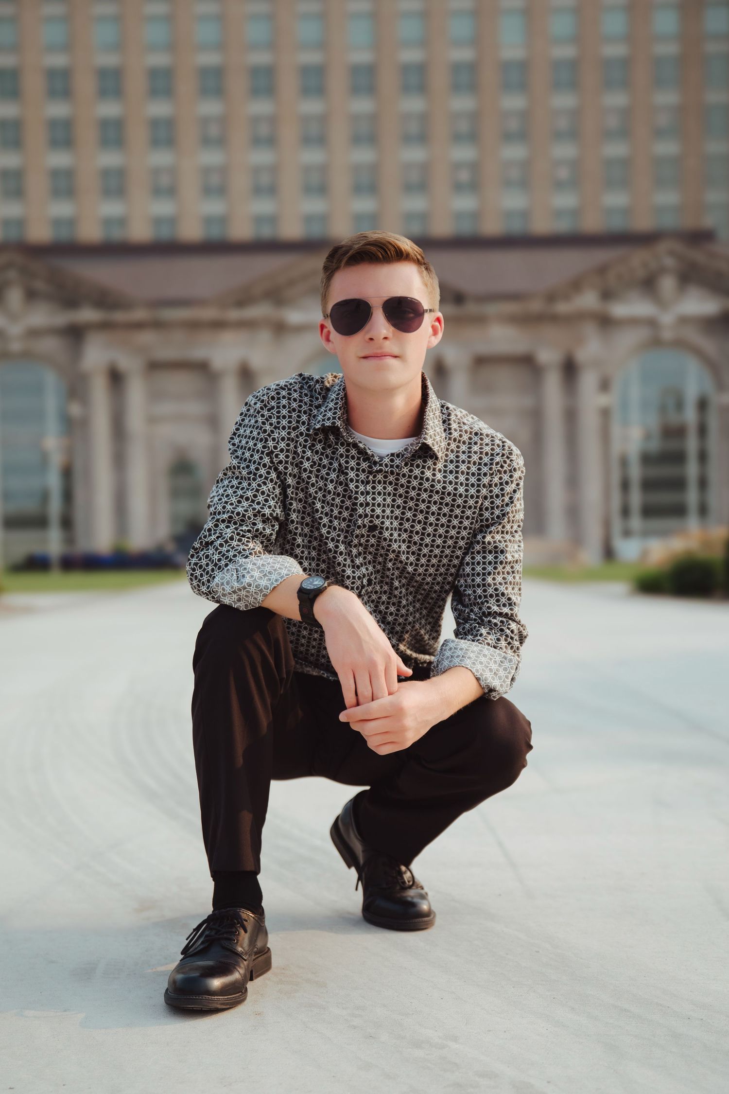 Stylish young man in patterned shirt and sunglasses crouches confidently in front of a large building.