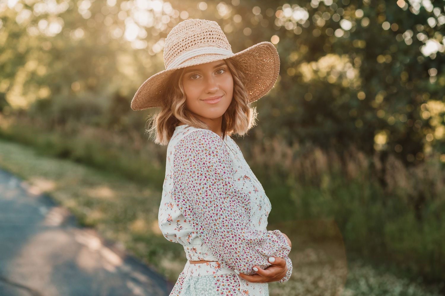Woman in floral dress and wide-brim straw hat smiling in warm golden hour sunlight.
