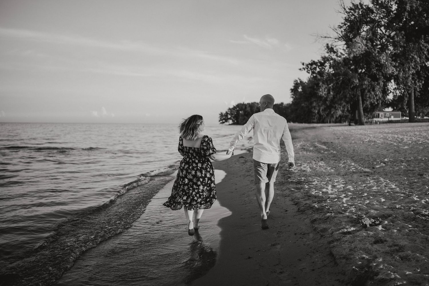 Black and white photo of a couple walking hand in hand along a scenic tree-lined beach.