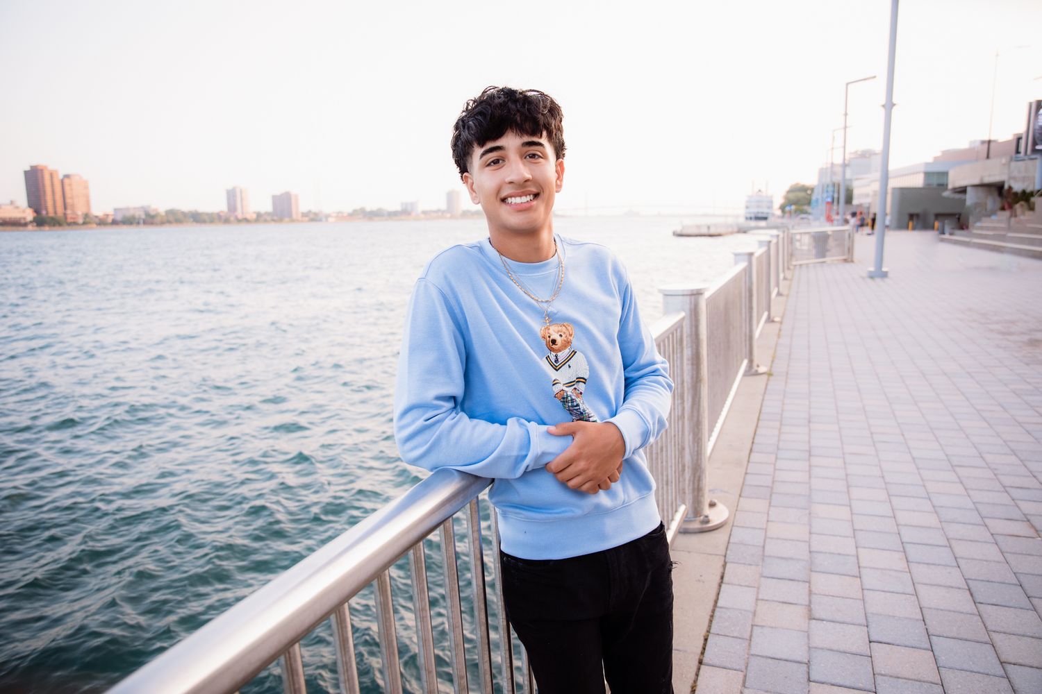 Smiling teen in blue sweatshirt poses on waterfront promenade with city skyline in background.