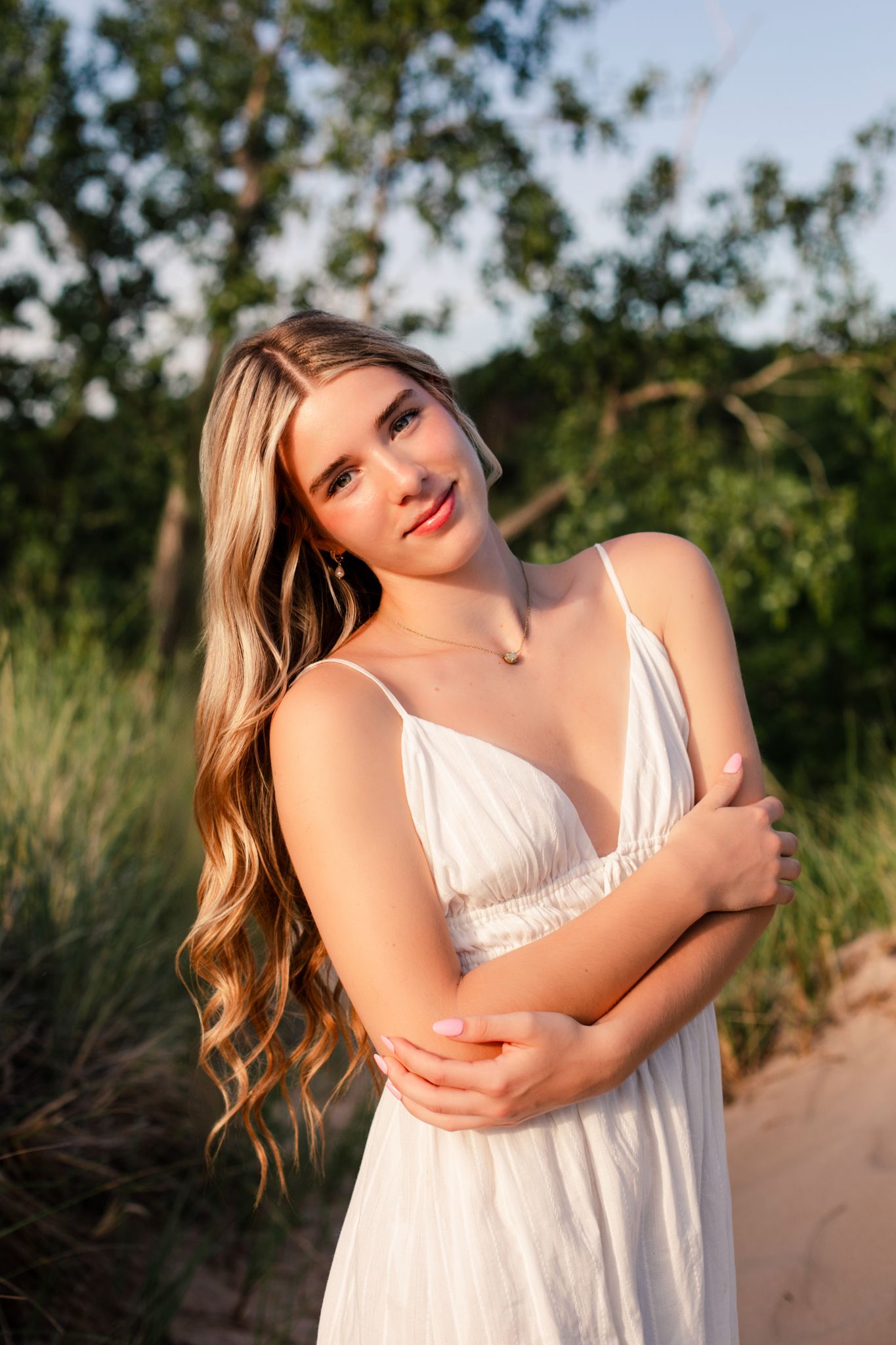 Young blonde woman in white spaghetti strap dress posing on sandy beach with arms crossed and soft smile.