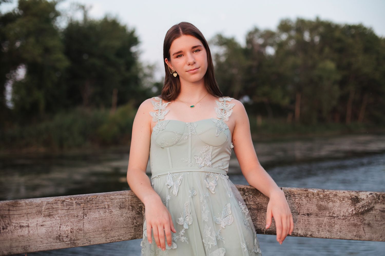 Teen girl in sage green lace dress leans against wooden railing outdoors at dusk.