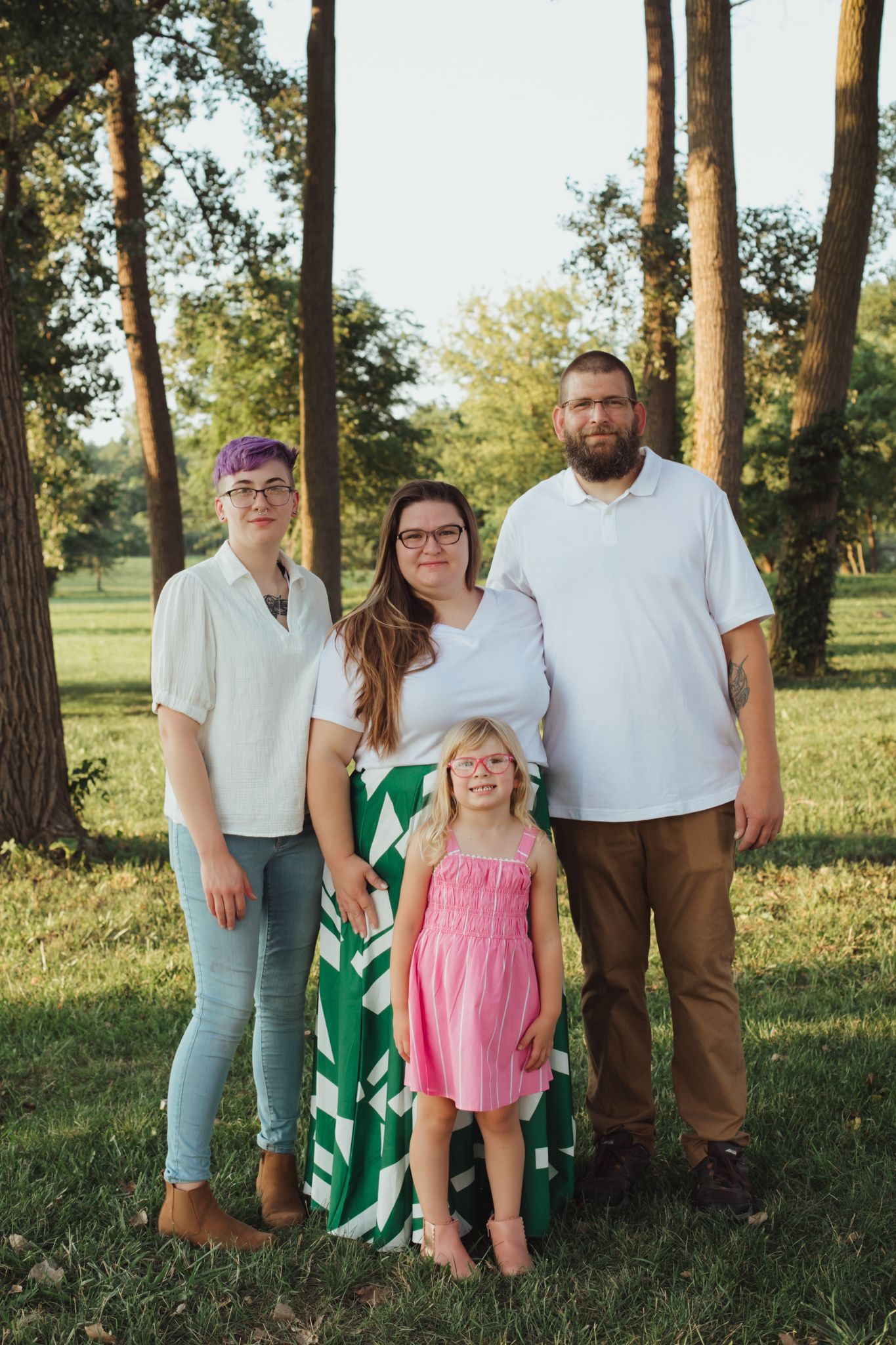 Family of four posing outdoors in a sunny park with tall trees, wearing casual summer outfits including white tops and a pink dress.