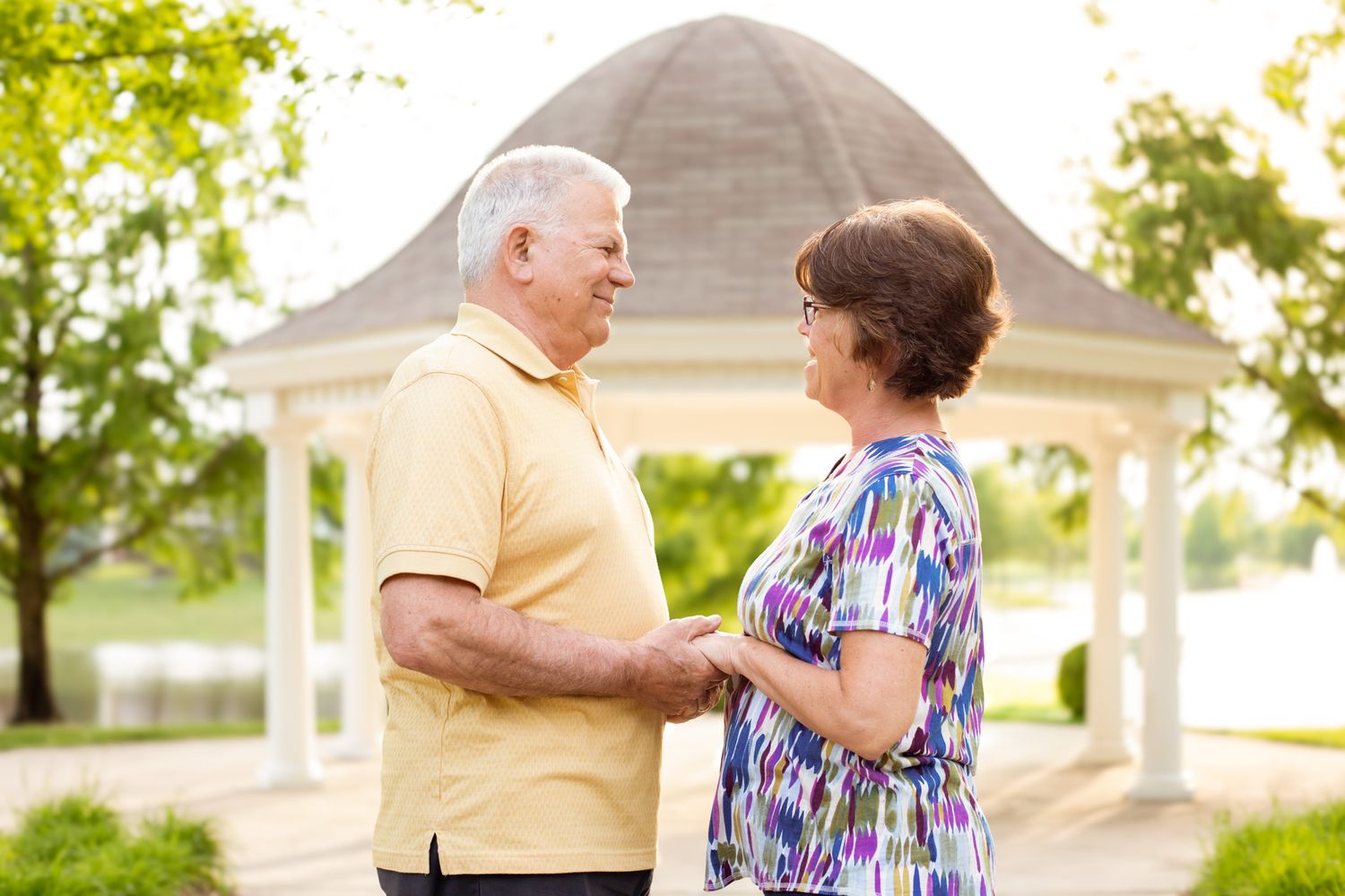 Senior couple holding hands and smiling at each other near a white gazebo in a lush green park.