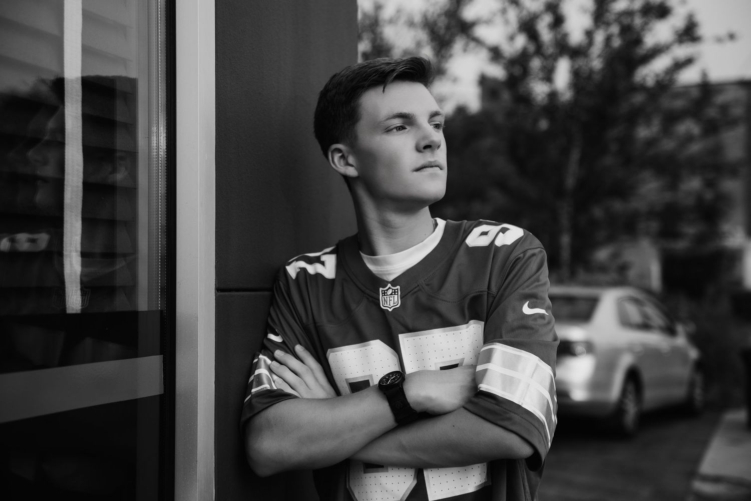 Young man in NFL jersey leaning against wall with arms crossed, black and white portrait photo.