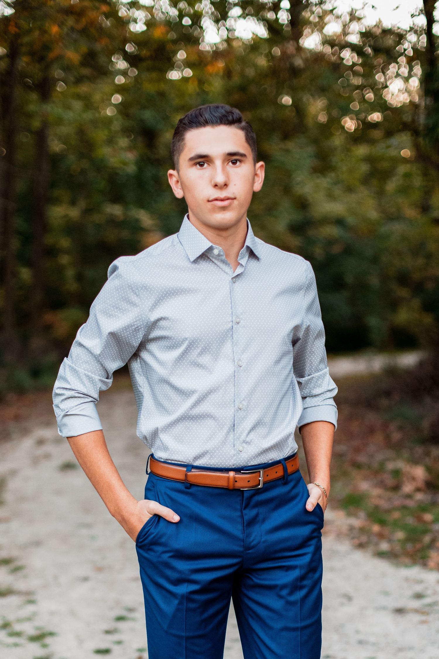 Young man in light blue shirt and navy pants with brown belt poses on a gravel path surrounded by trees.