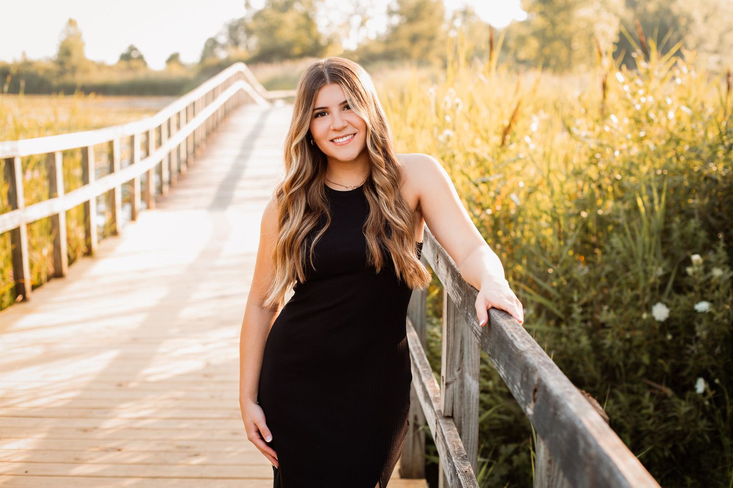 Smiling woman in black dress leaning on railing of sunny boardwalk in golden field.
