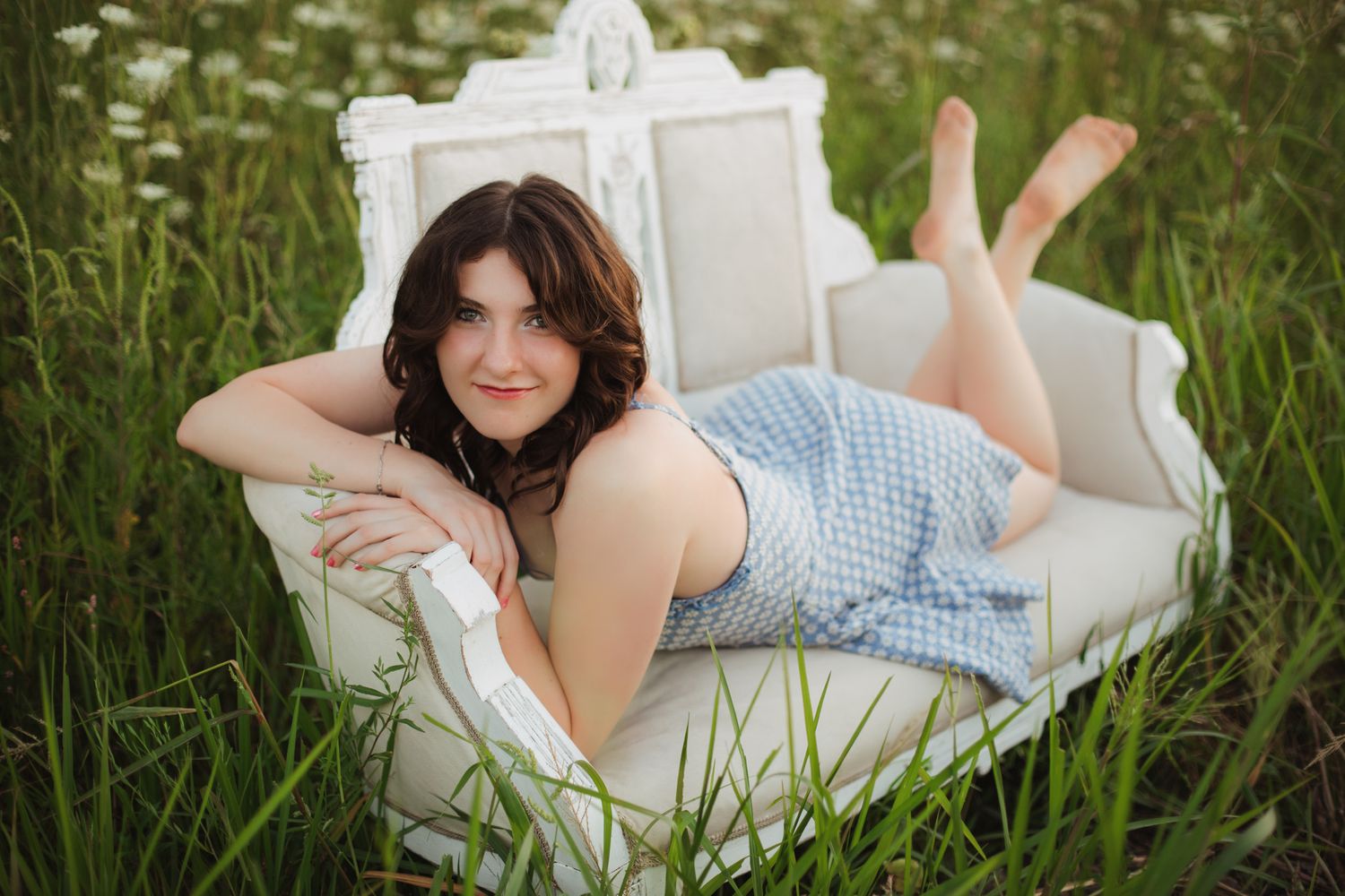 Smiling young woman in blue gingham dress lying on white ornate sofa in green meadow.