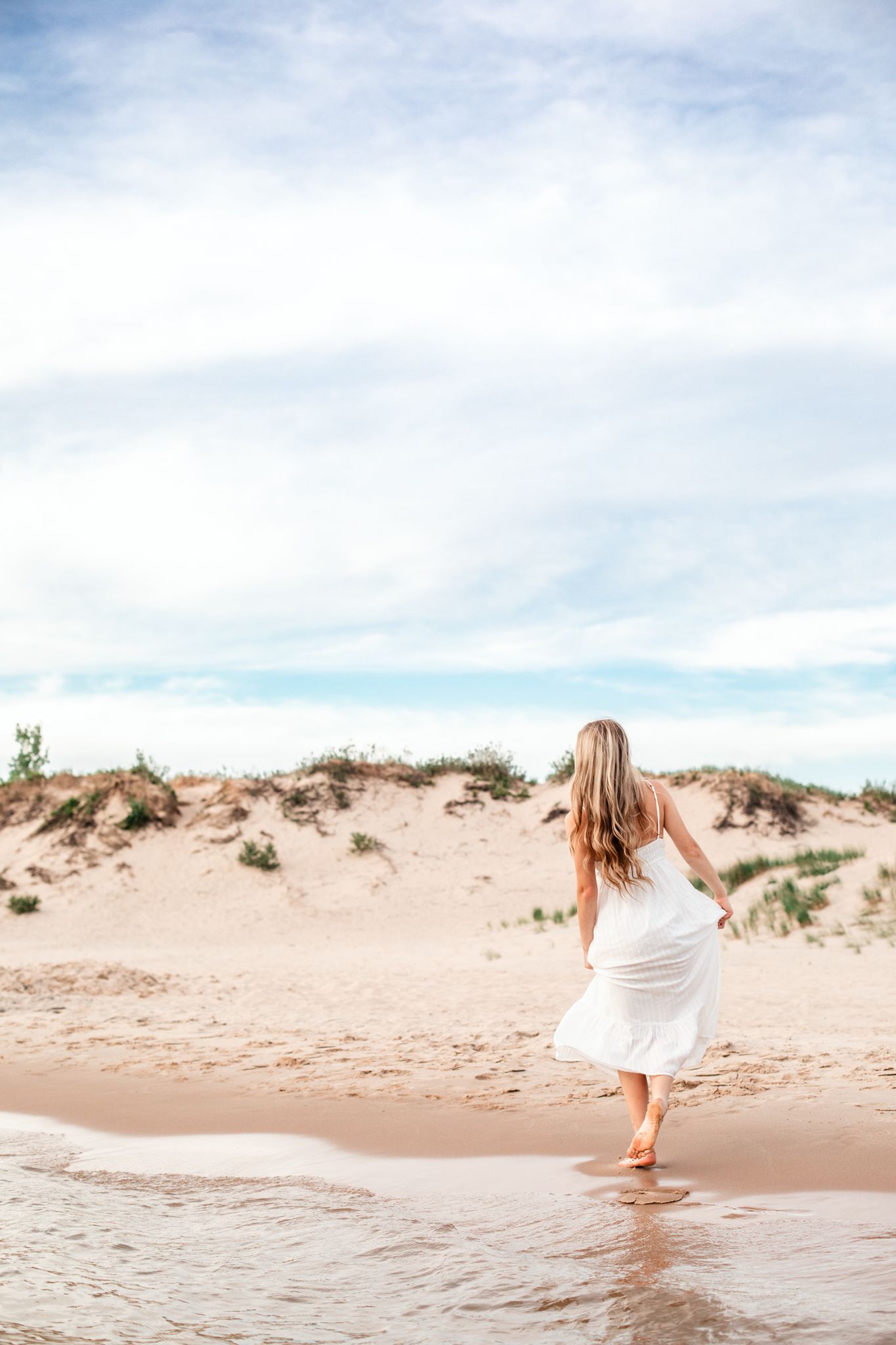 Barefoot woman in flowing white dress walking along sandy beach toward sand dunes.