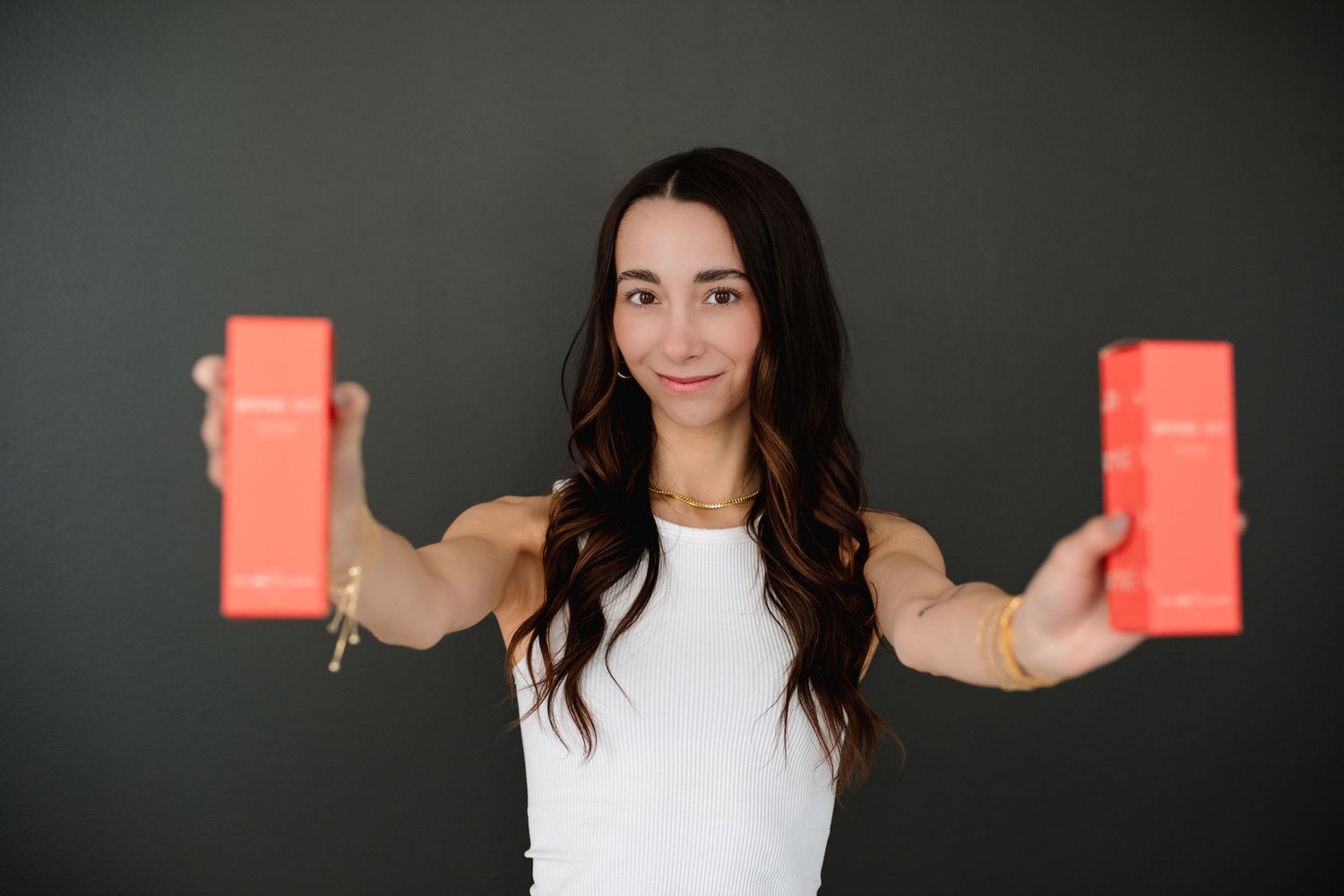 Smiling brunette woman in white tank top holding out two coral Mane Ivy product boxes toward camera.