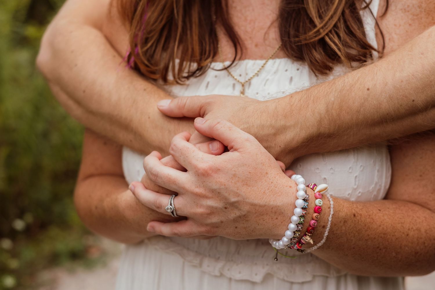 Couple embracing, woman in white dress with colorful beaded bracelets and ring, hands clasped together in romantic outdoor setting.