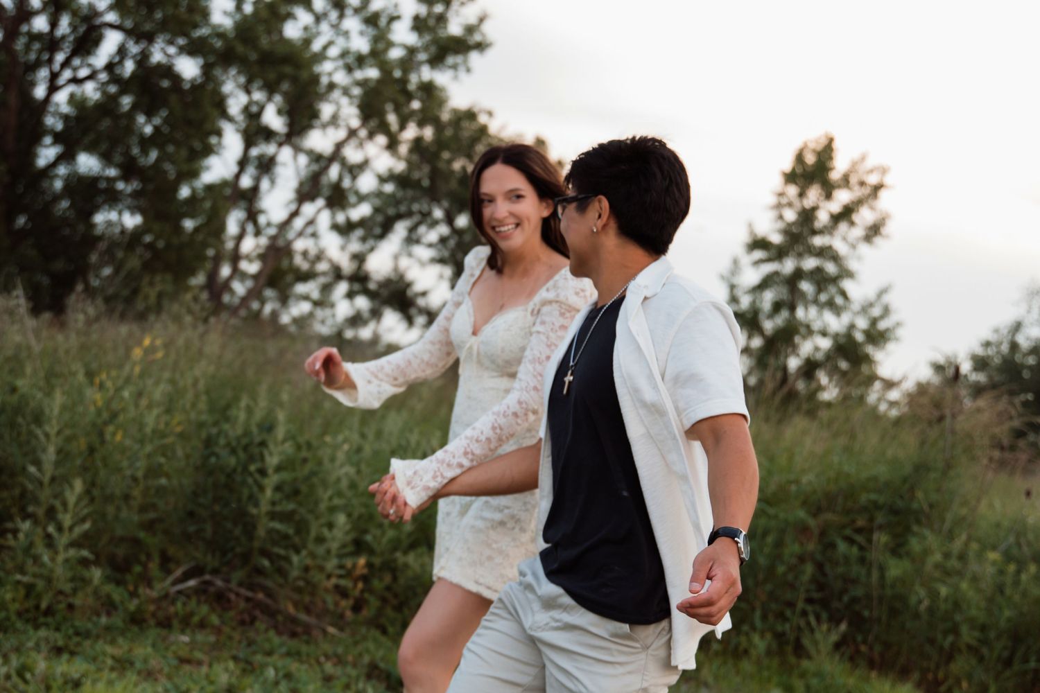 A happy couple walks hand-in-hand through a scenic green field during a warm sunset.