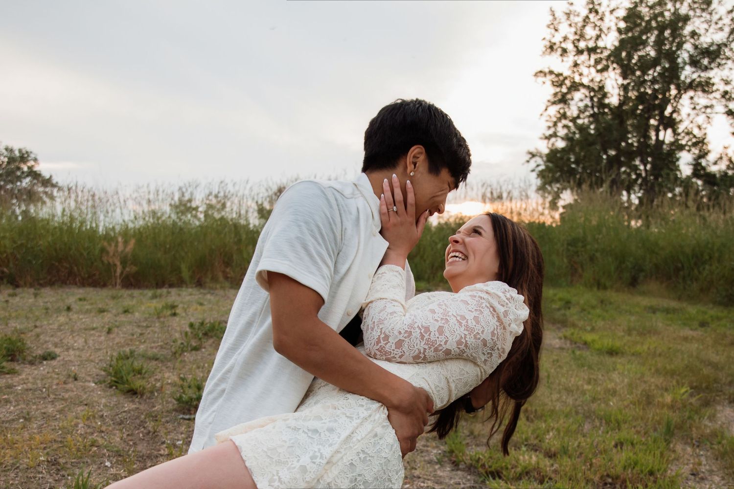 A couple shares a romantic moment outdoors, the woman smiling as her partner leans in close against a lush green backdrop.