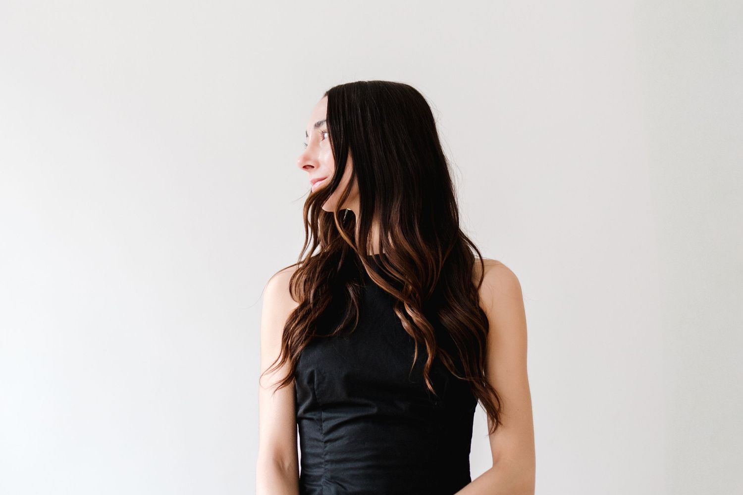Woman with long dark wavy hair wearing black dress photographed from behind against white wall.