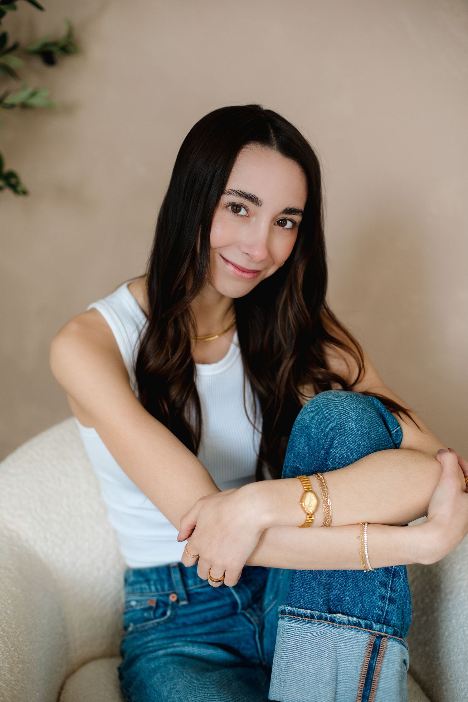 Dark-haired woman in white tank top and jeans sitting in white chair with gold bracelet and watch.