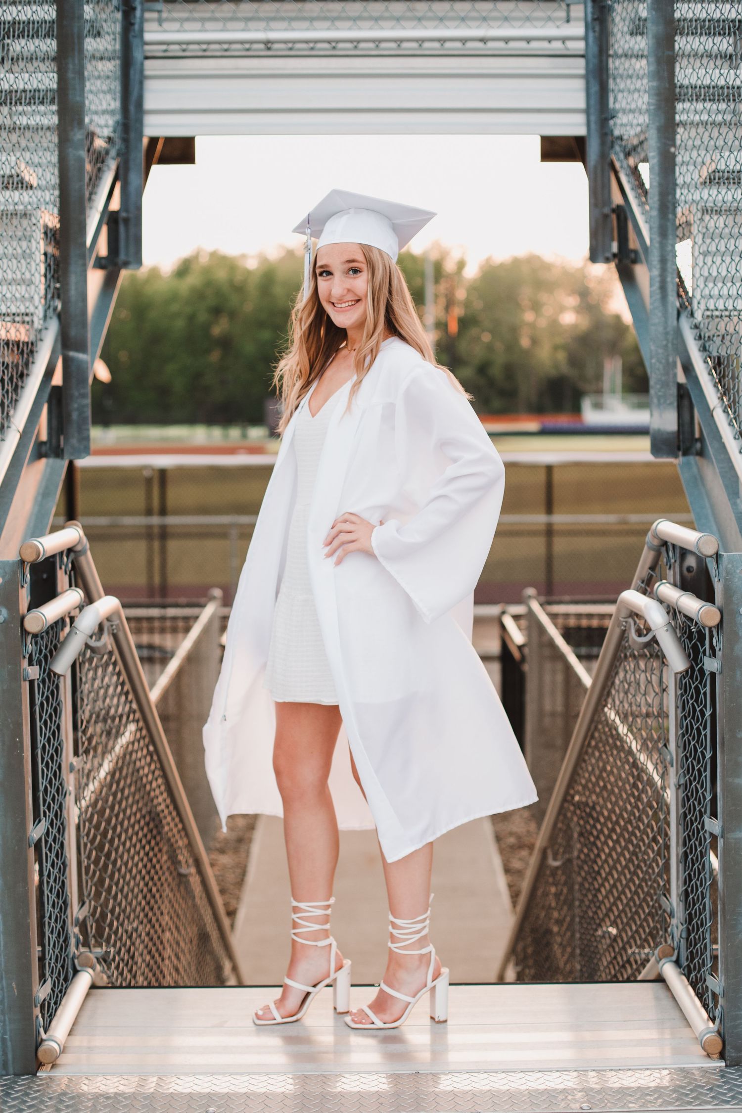 Smiling graduate poses in white cap and gown on stadium stairs wearing strappy heeled sandals.