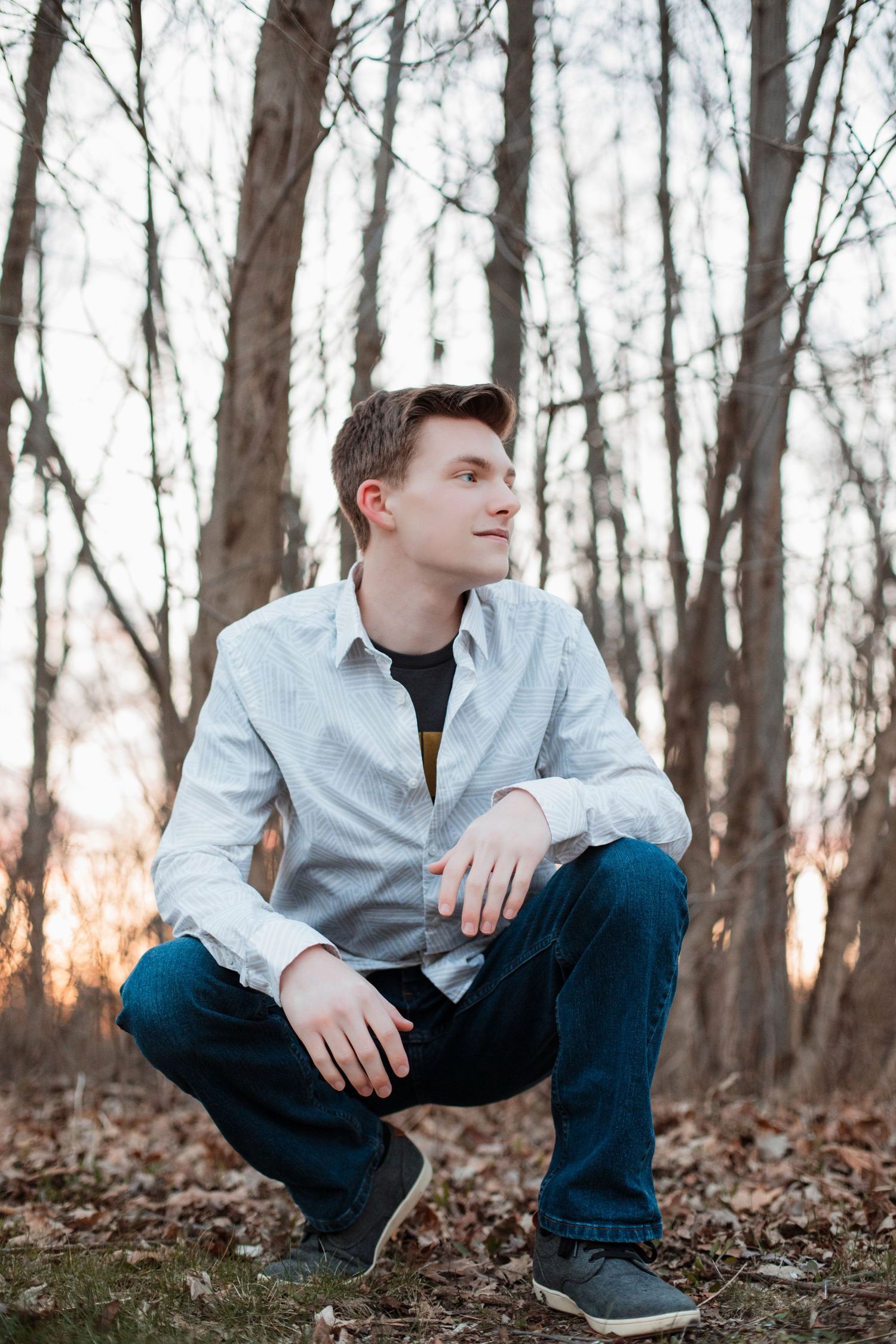 Thoughtful teen boy in white button-up shirt crouches and gazes upward in a bare autumn forest.