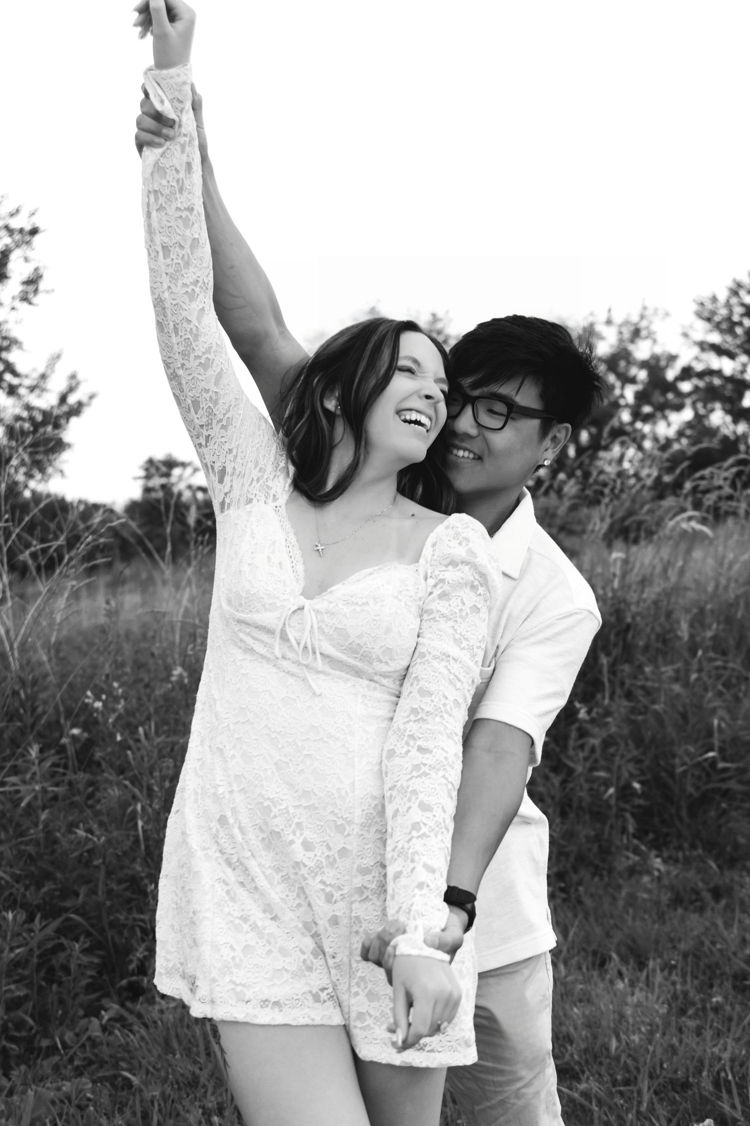 A joyful couple dances in a field, woman in white dress with arm raised, in black and white photo.