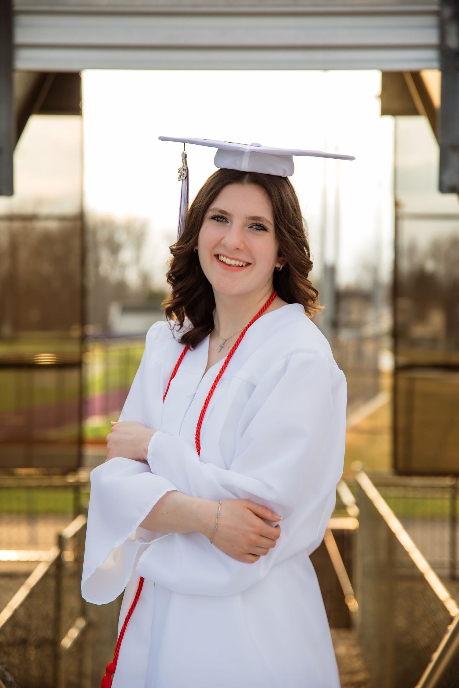 Smiling young woman in white graduation cap and gown with red trim, arms crossed, posing outdoors at sunset.