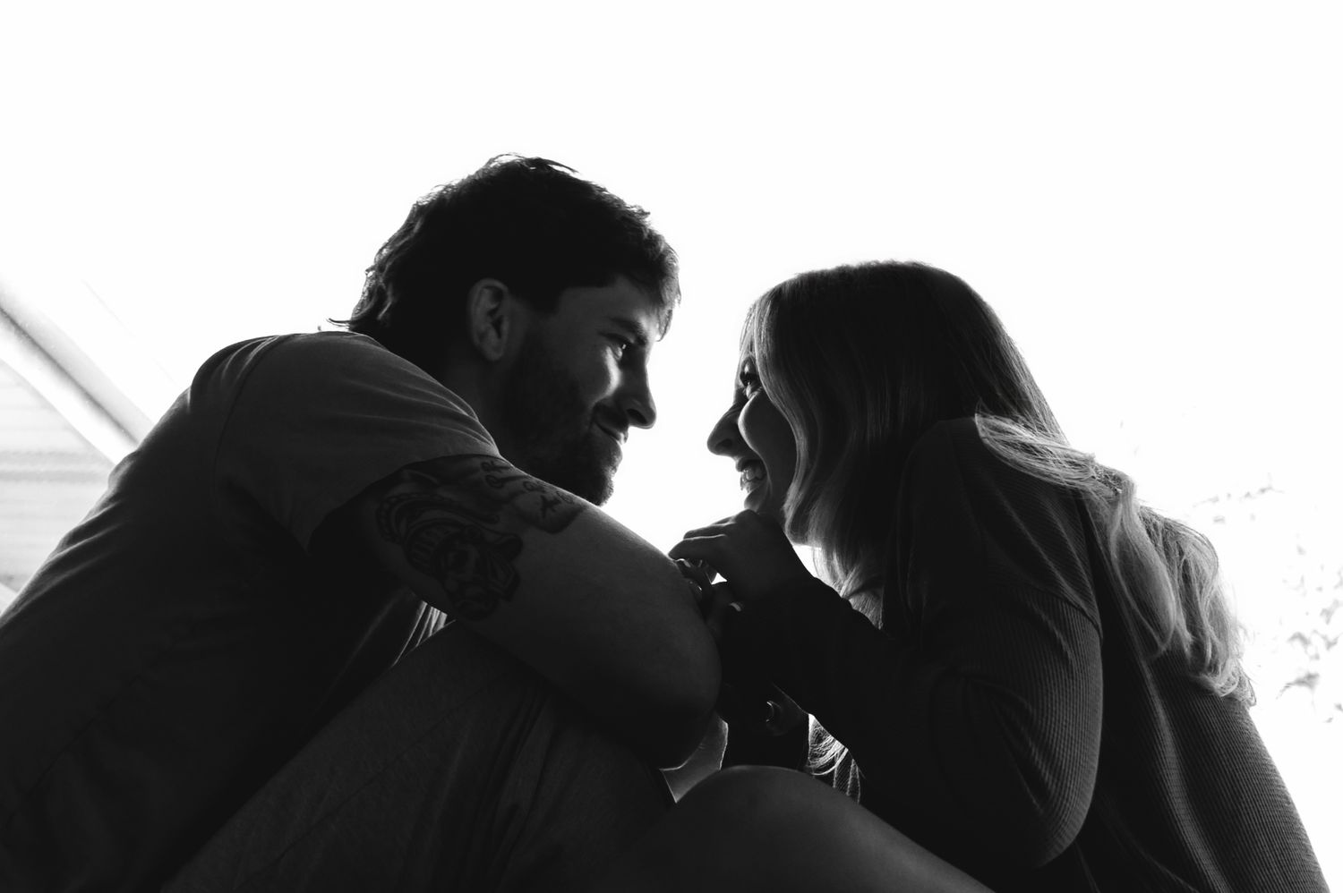 Black and white photo of a couple sharing an intimate moment, smiling and gazing at each other in soft natural light.