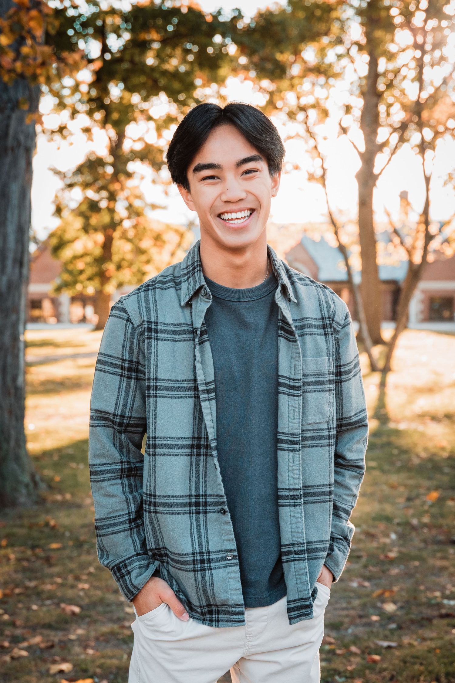 Smiling young Asian man in plaid shirt and white pants standing outdoors in a park during golden hour.