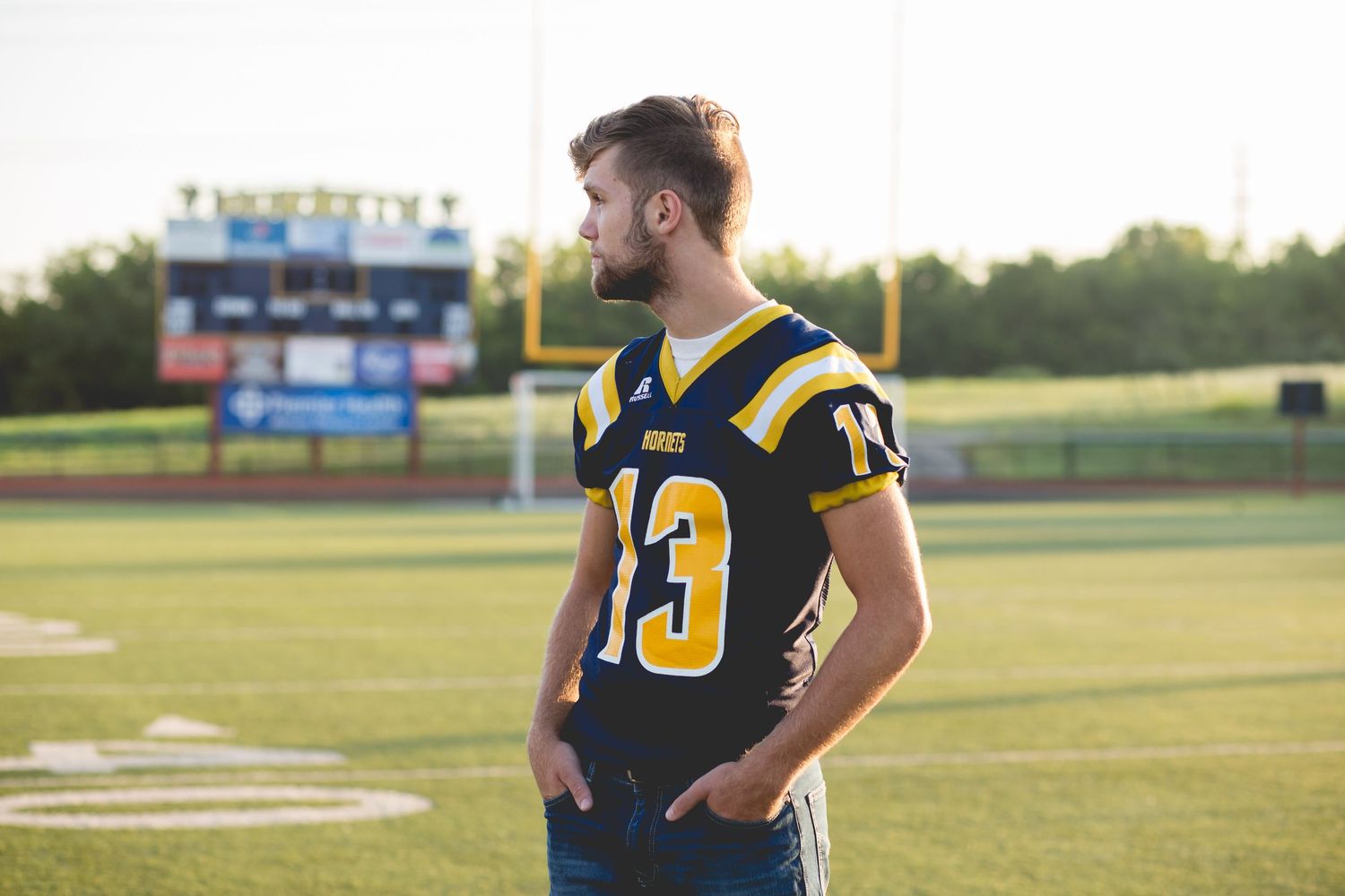 Young man in navy and gold football jersey number 13 stands on green field near stadium bleachers.