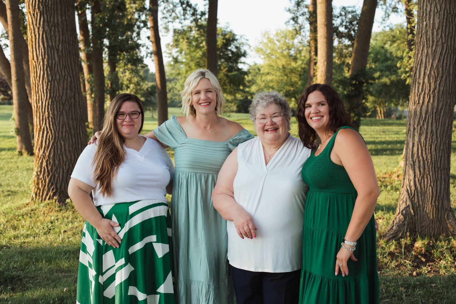 Four women posing together outdoors in a park, wearing green and white outfits, smiling warmly.