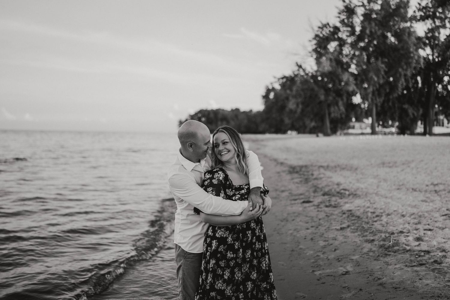 Black and white image of a couple sharing a tender embrace on a sandy beach by the water.