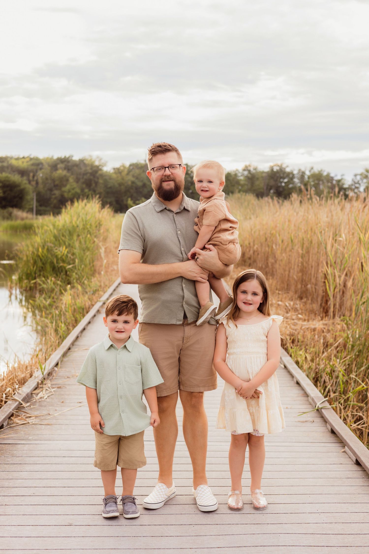 A bearded father holds a baby while standing on a wooden boardwalk with two young children in neutral outdoor attire.