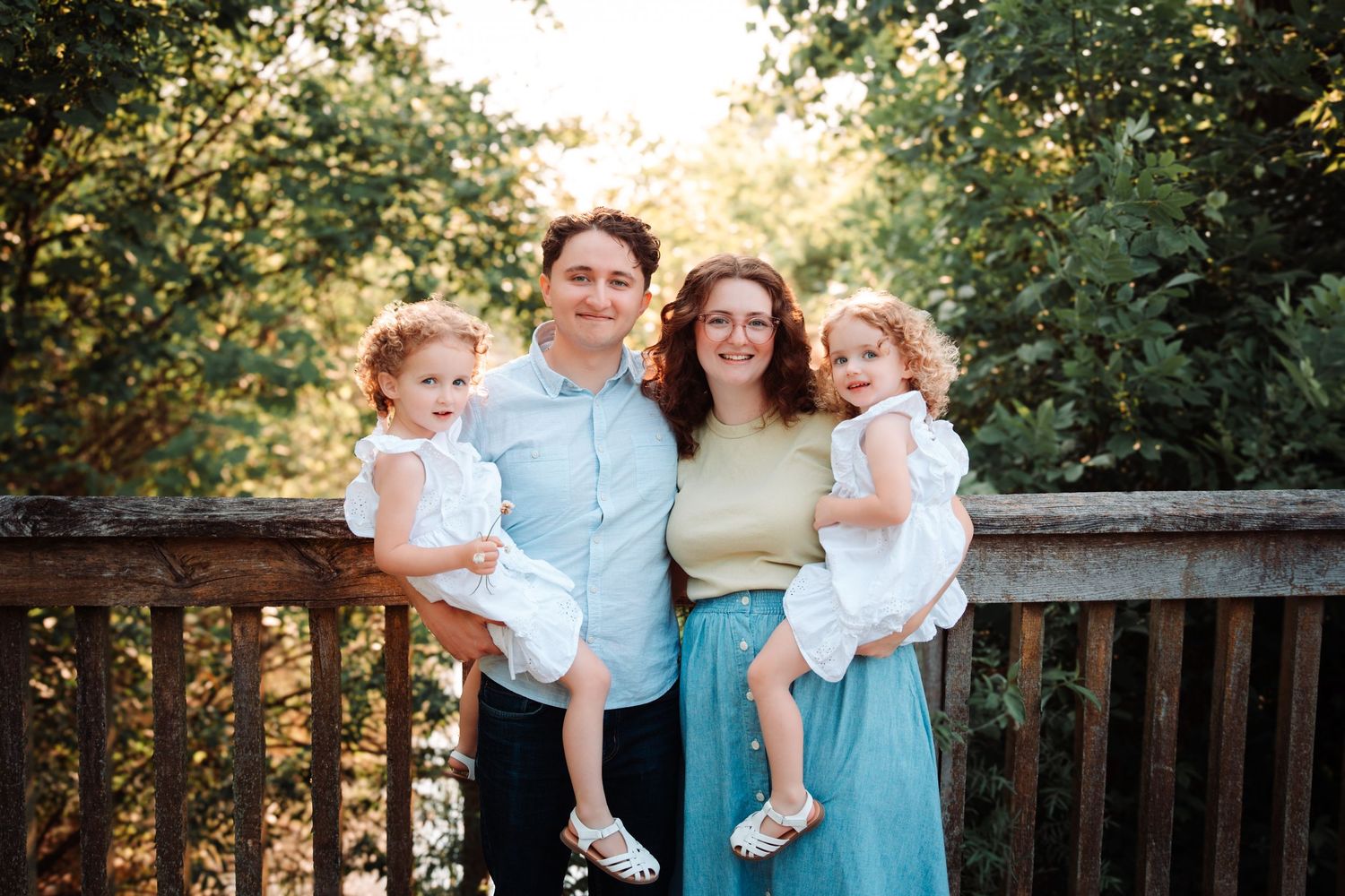 A happy couple holding their two young daughters in white dresses while standing on a wooden bridge outdoors.