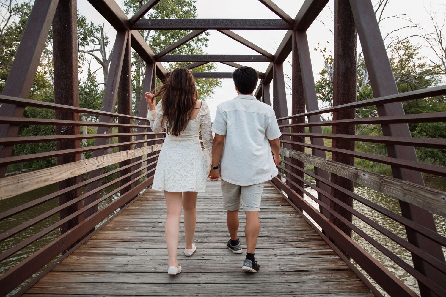 Couple holding hands walking across rustic wooden bridge surrounded by trees.