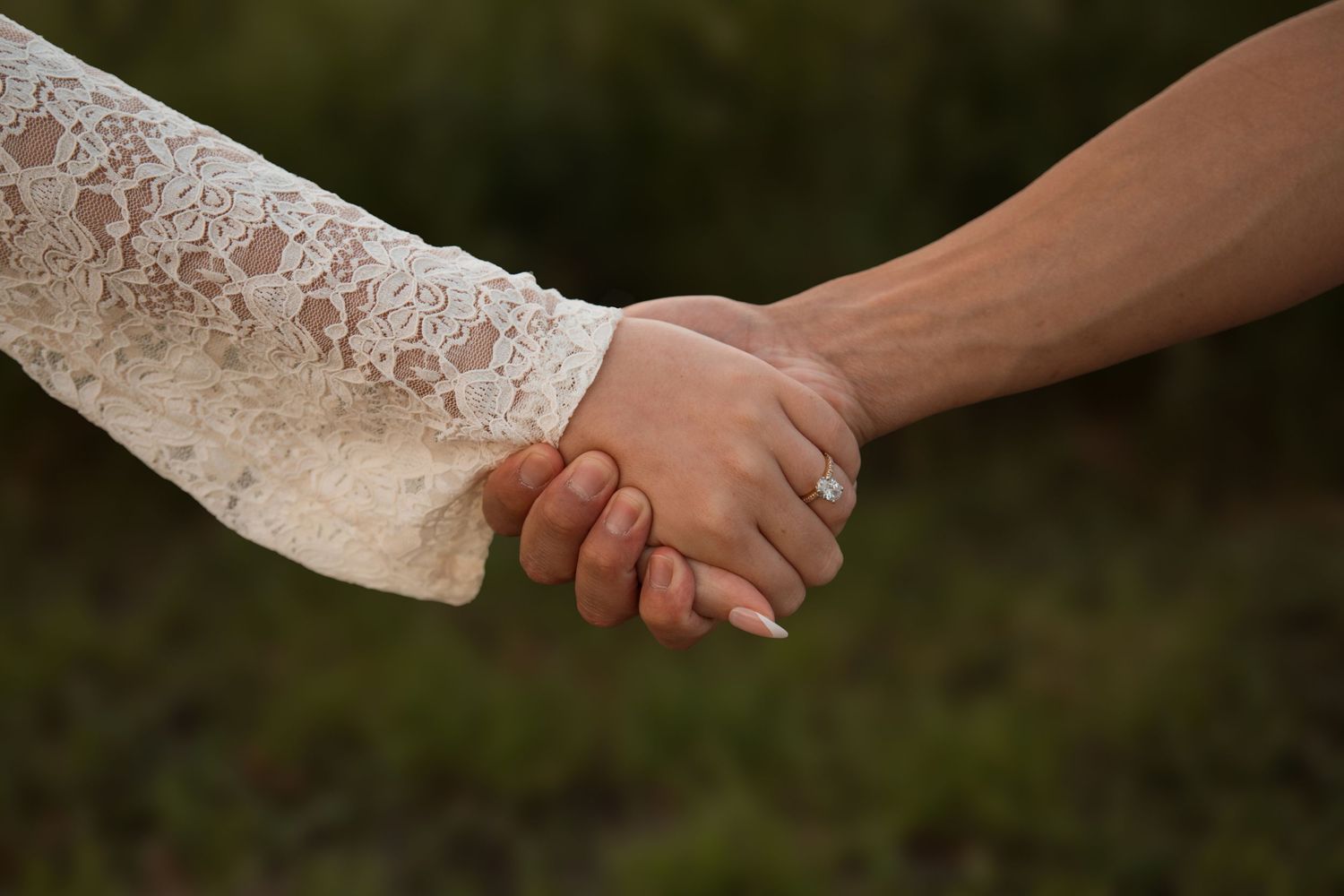 Close-up of couple holding hands, woman wearing white lace sleeve and diamond engagement ring outdoors.