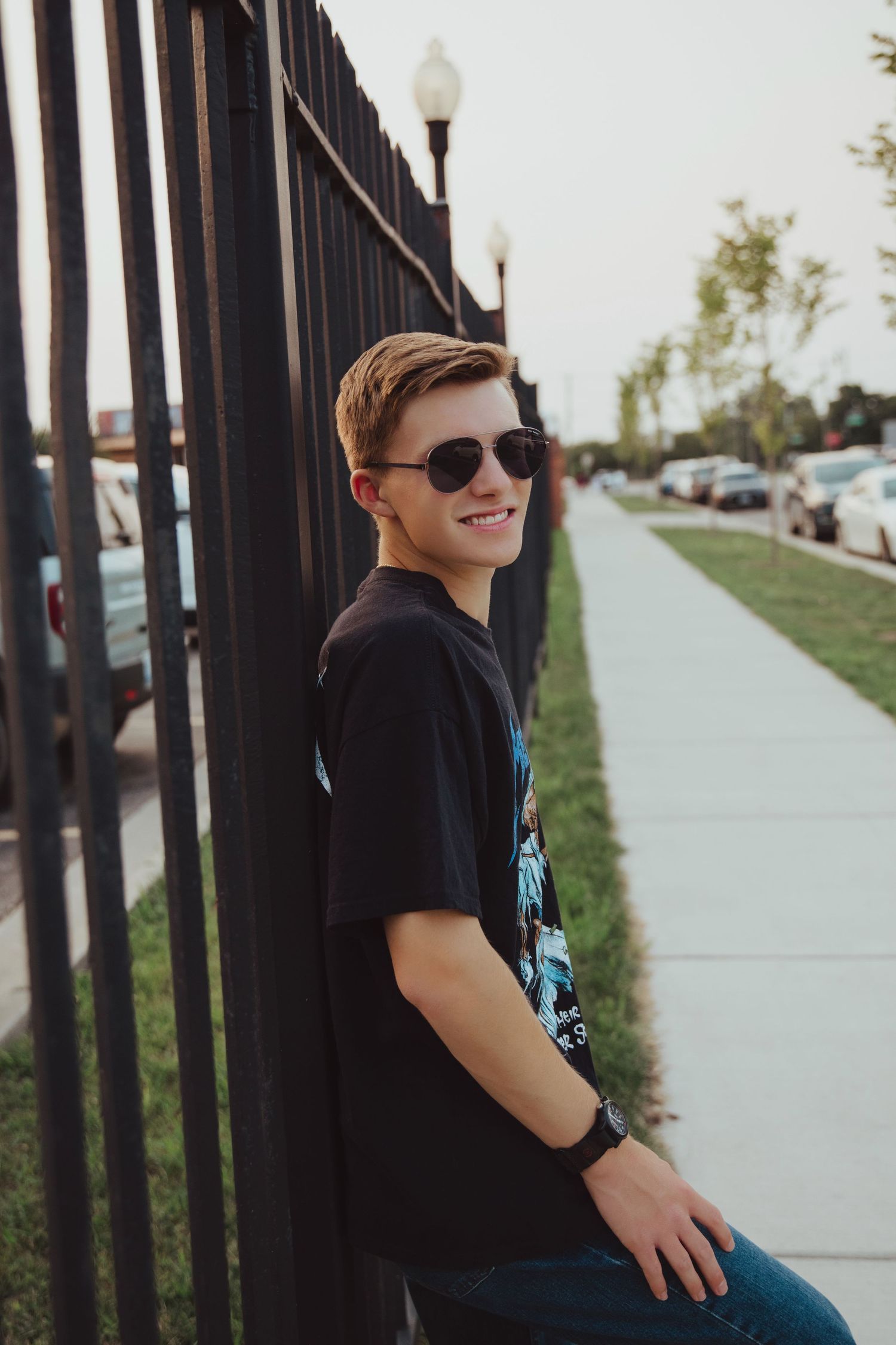 Smiling teen in black graphic tee and sunglasses leaning against iron fence on sidewalk.