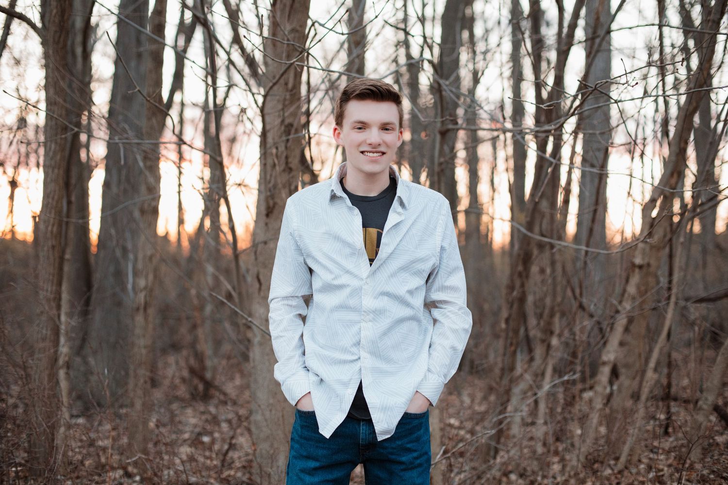 Smiling young man in light gray jacket stands with hands in pockets in a bare winter forest.