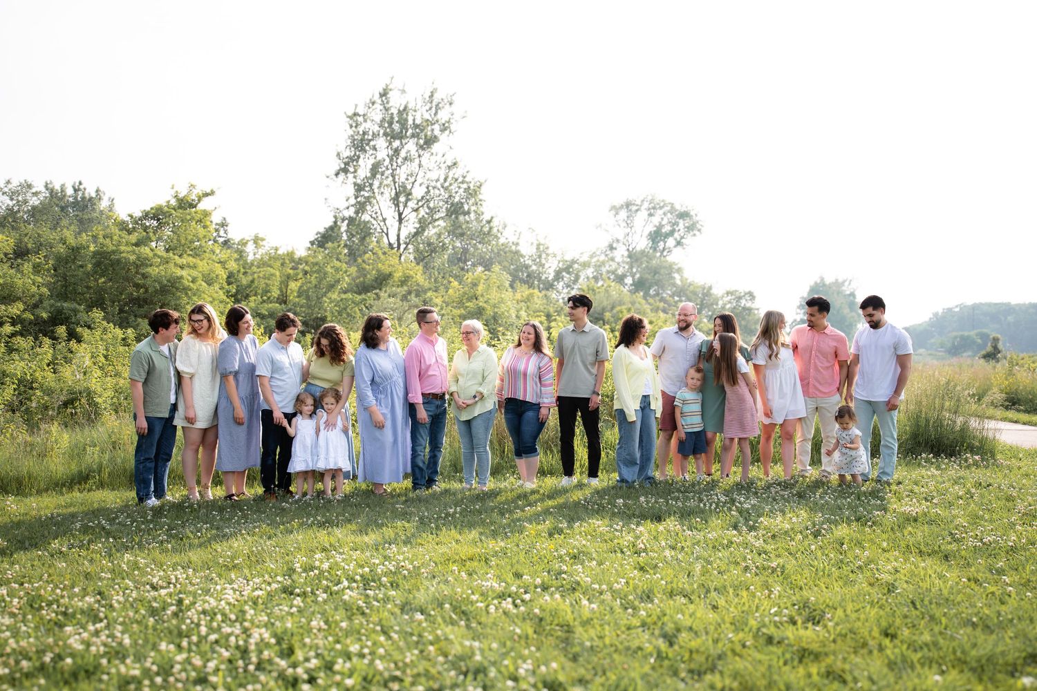 Large family holding hands in a line across a sunny wildflower meadow with trees behind.