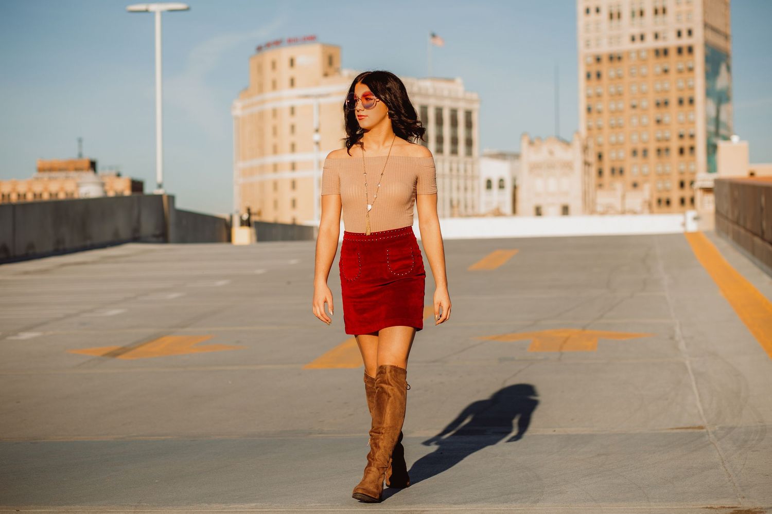 Young woman in red mini skirt and brown boots walking on urban rooftop parking deck with city buildings behind her.