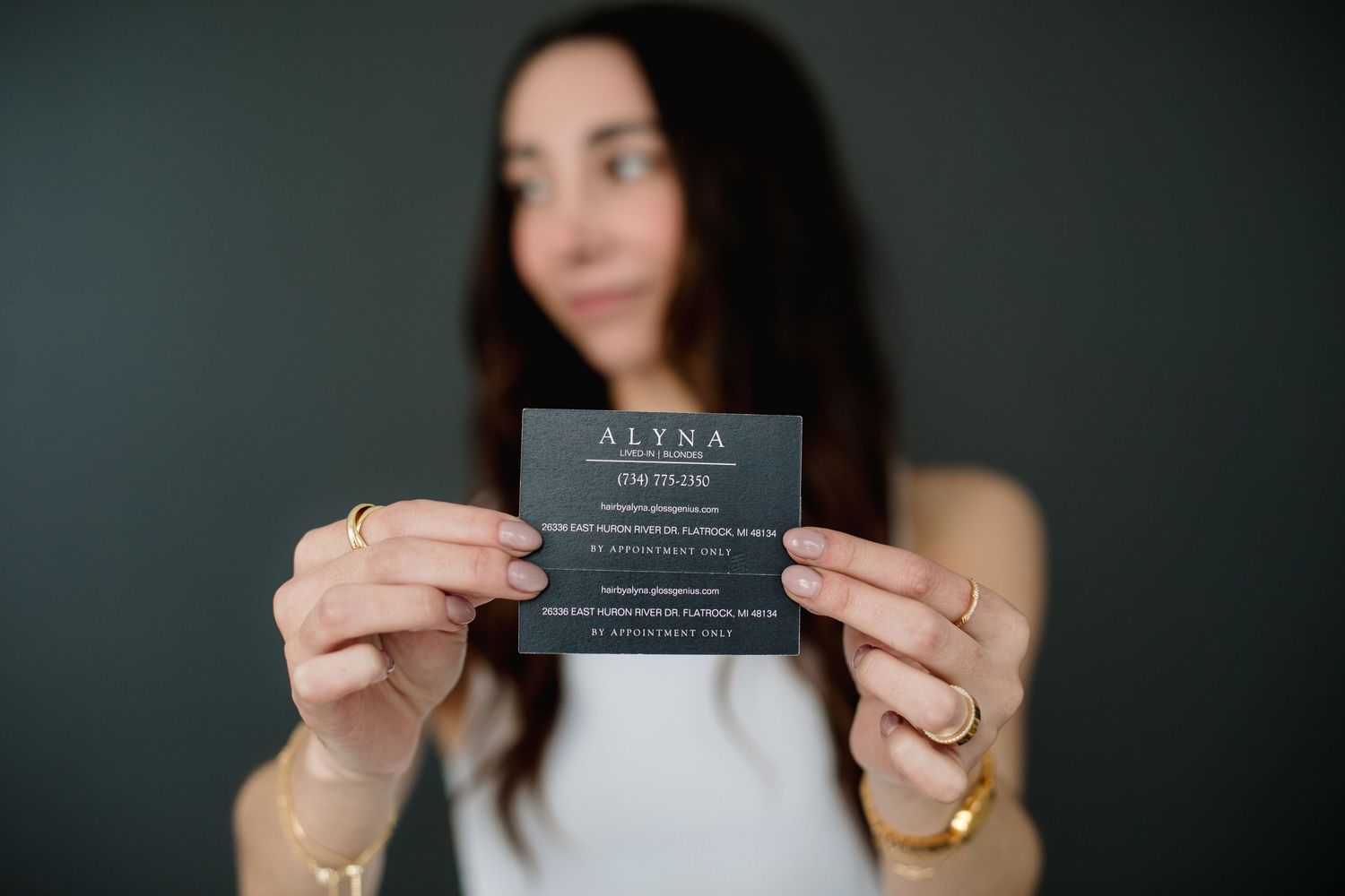 Woman holding a dark business card for Alyna hair salon toward the camera.