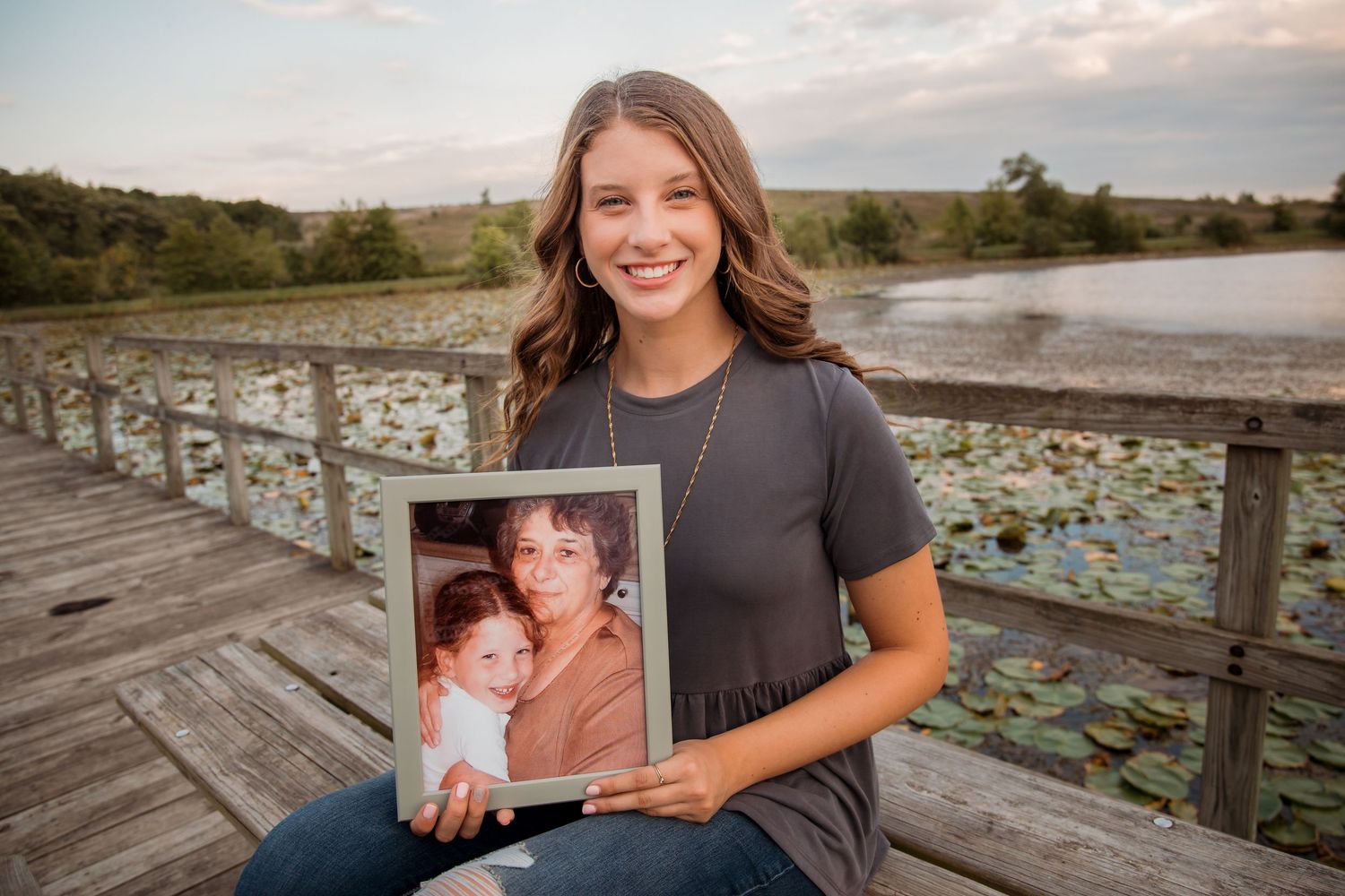 Smiling teen girl holding framed childhood photo on wooden dock near lily pad pond.