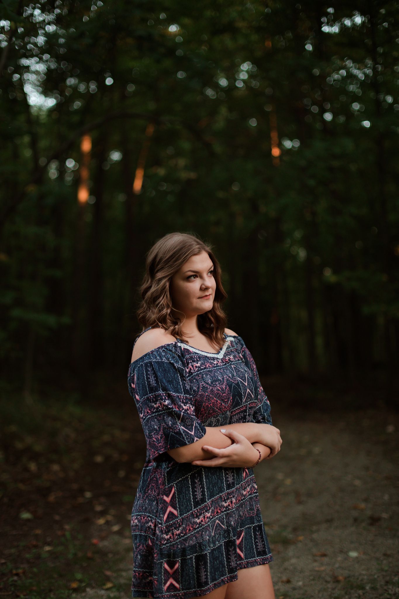 Young woman in patterned off-shoulder dress stands on forest path at dusk, arms crossed, looking over shoulder.