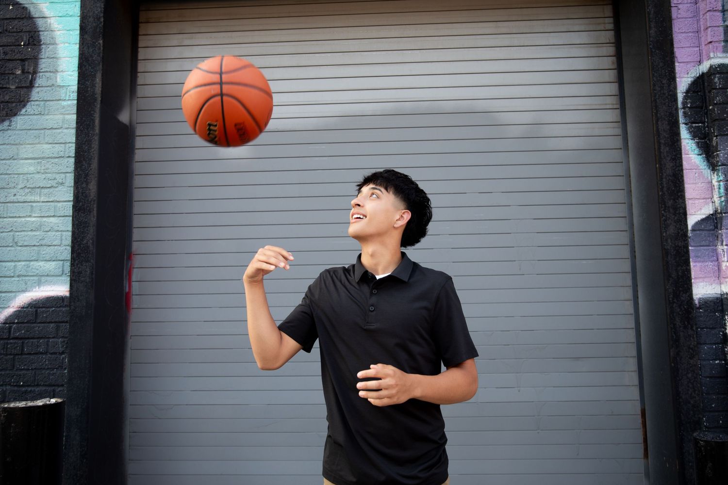 Young man in black outfit spinning a basketball on his finger outdoors near a graffiti wall.