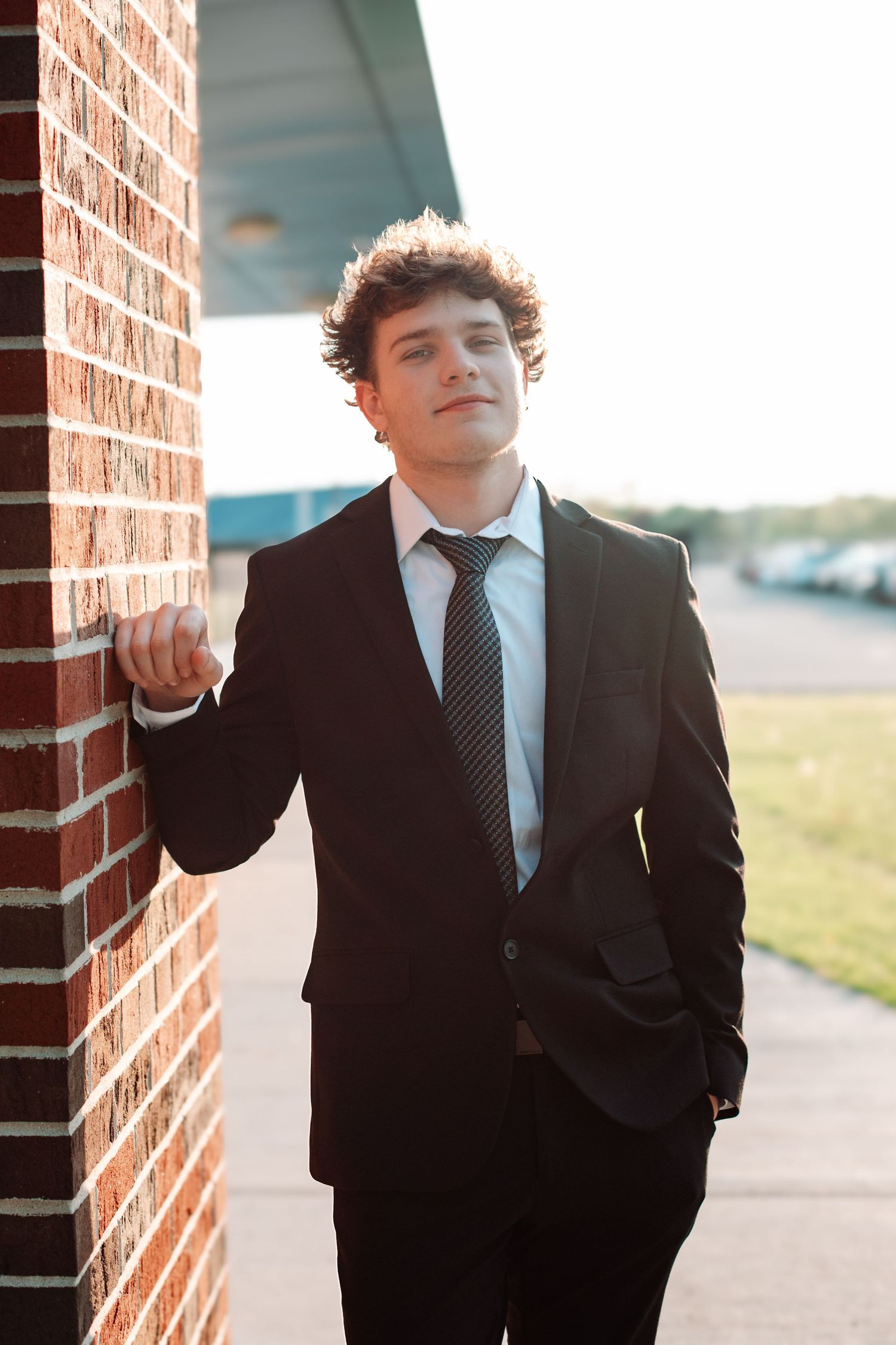 Young man in black suit and tie leaning against brick wall outdoors, posing confidently for a portrait photo.