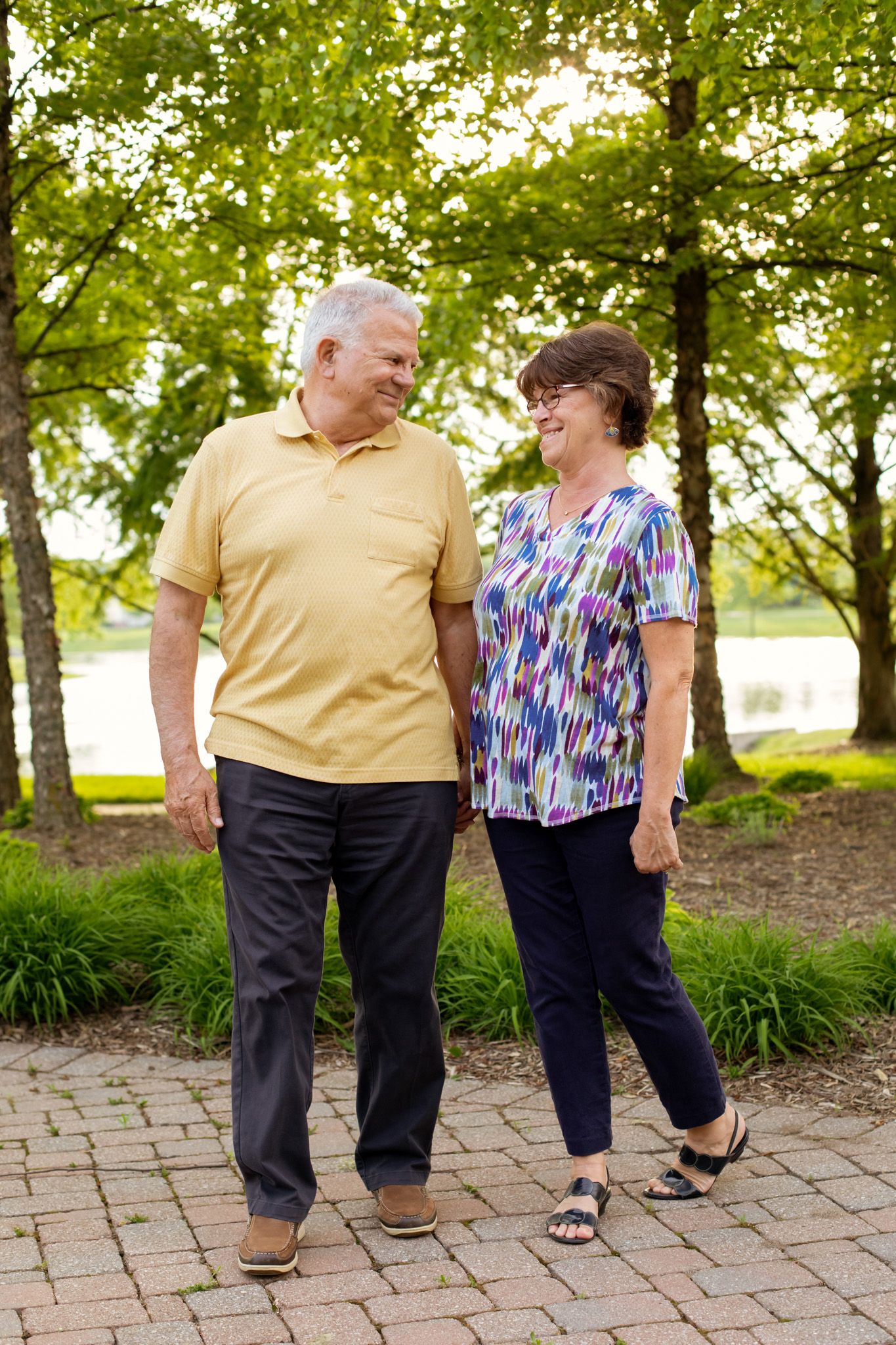 Senior couple walking hand in hand on a brick path in a lush green park, smiling at each other.