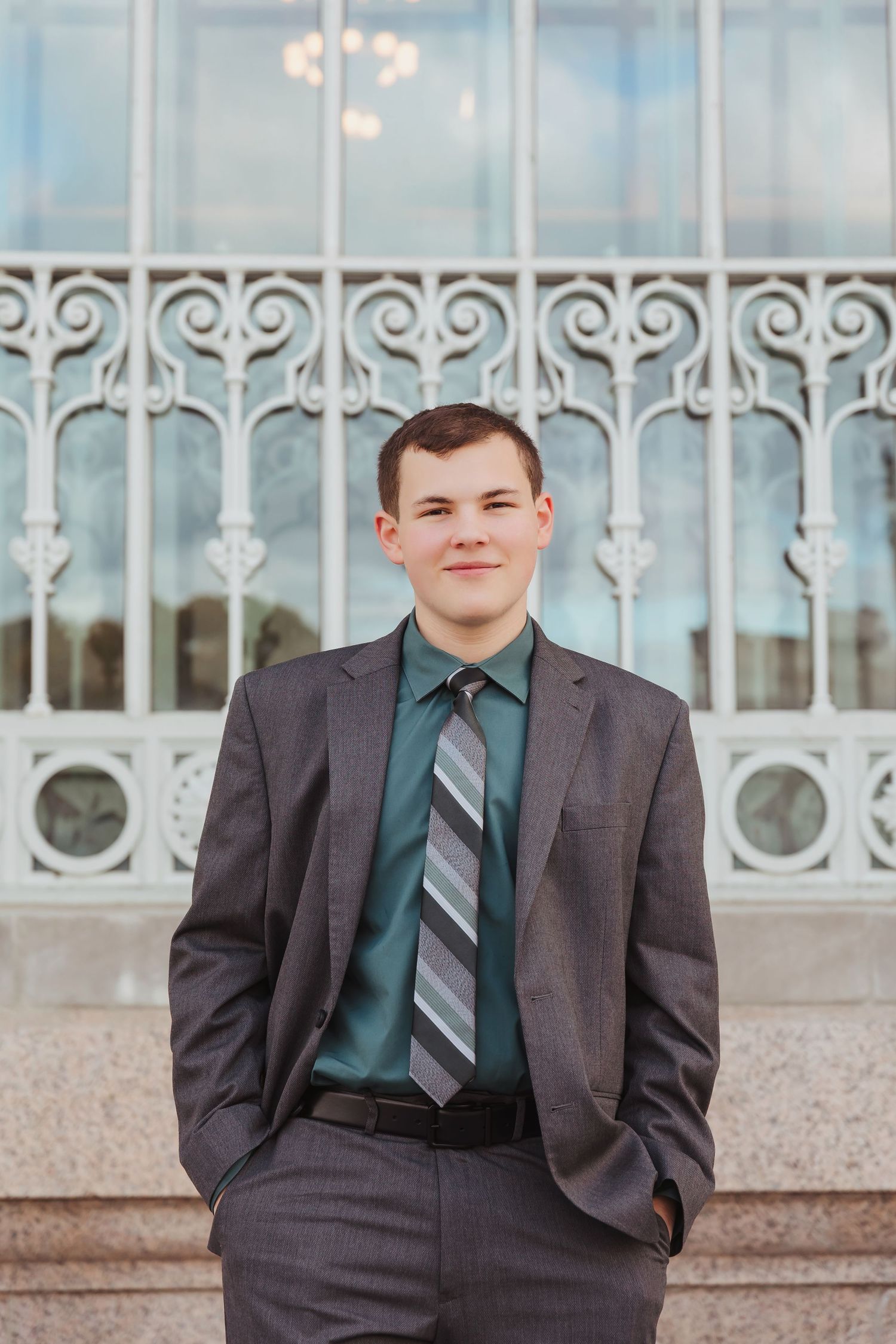 Young man in gray suit and striped tie smiling outdoors near ornate white iron gate.