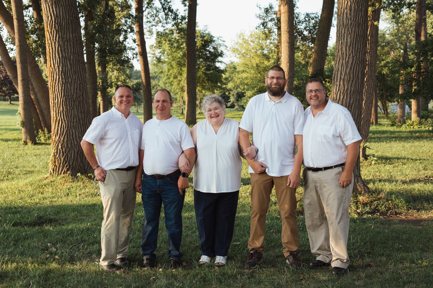 Family of five posing together outdoors in white shirts among tall trees on a sunny day.
