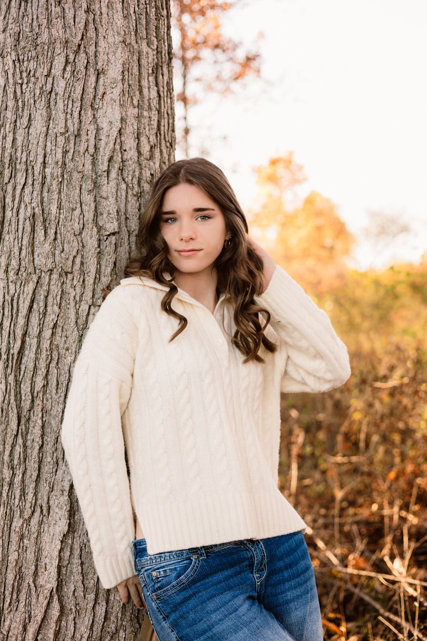 Young woman in cream cable-knit sweater and jeans leaning against a tree in an autumn outdoor setting.