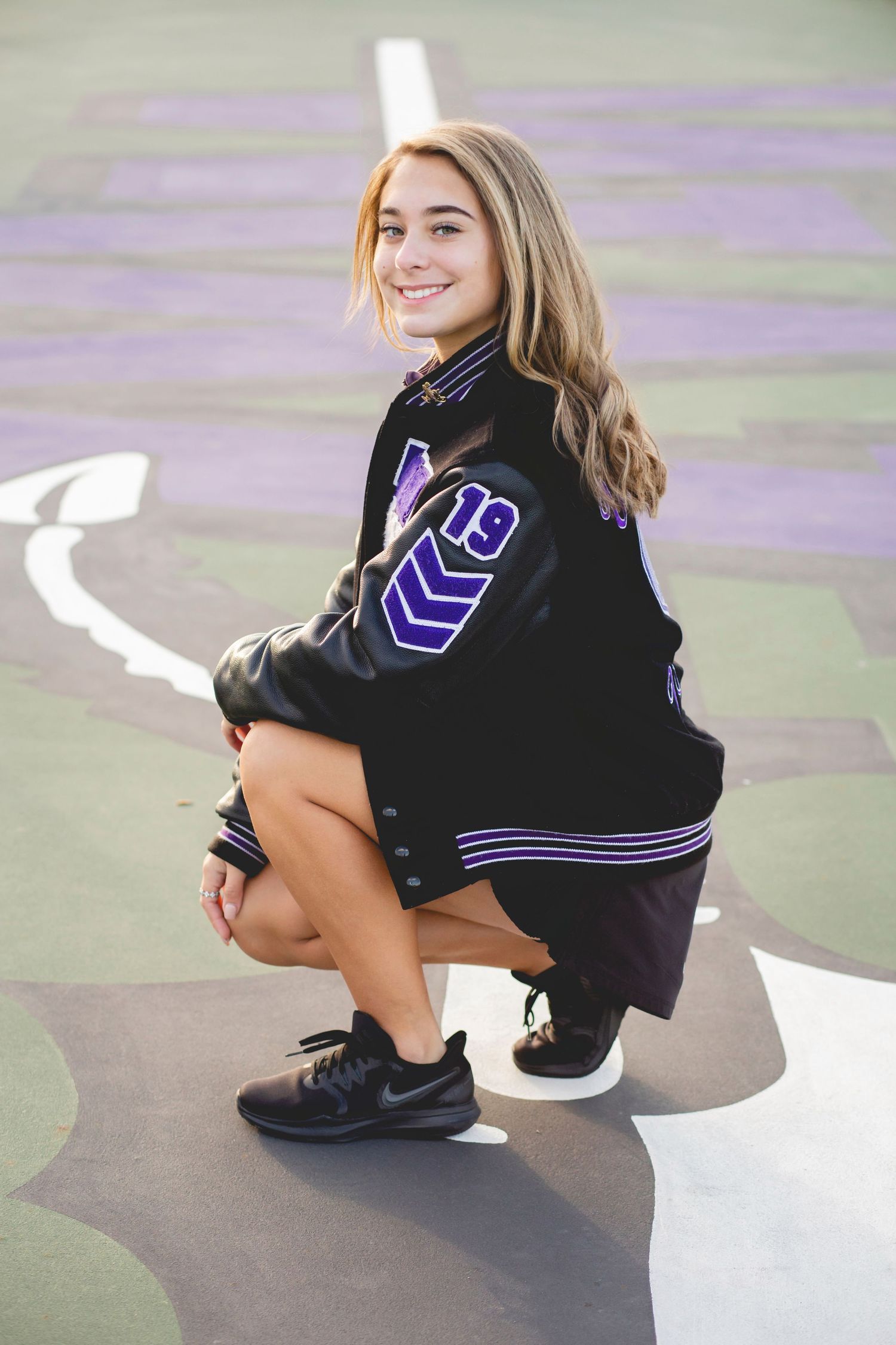 Teen girl in black and purple varsity jacket crouching on a painted court, smiling, wearing black sneakers.