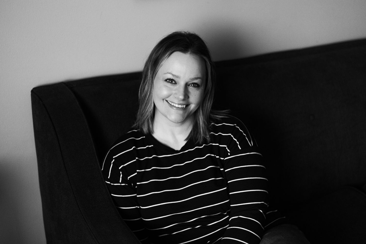 Cheerful woman in striped shirt smiling while seated on dark sofa in black and white photo.